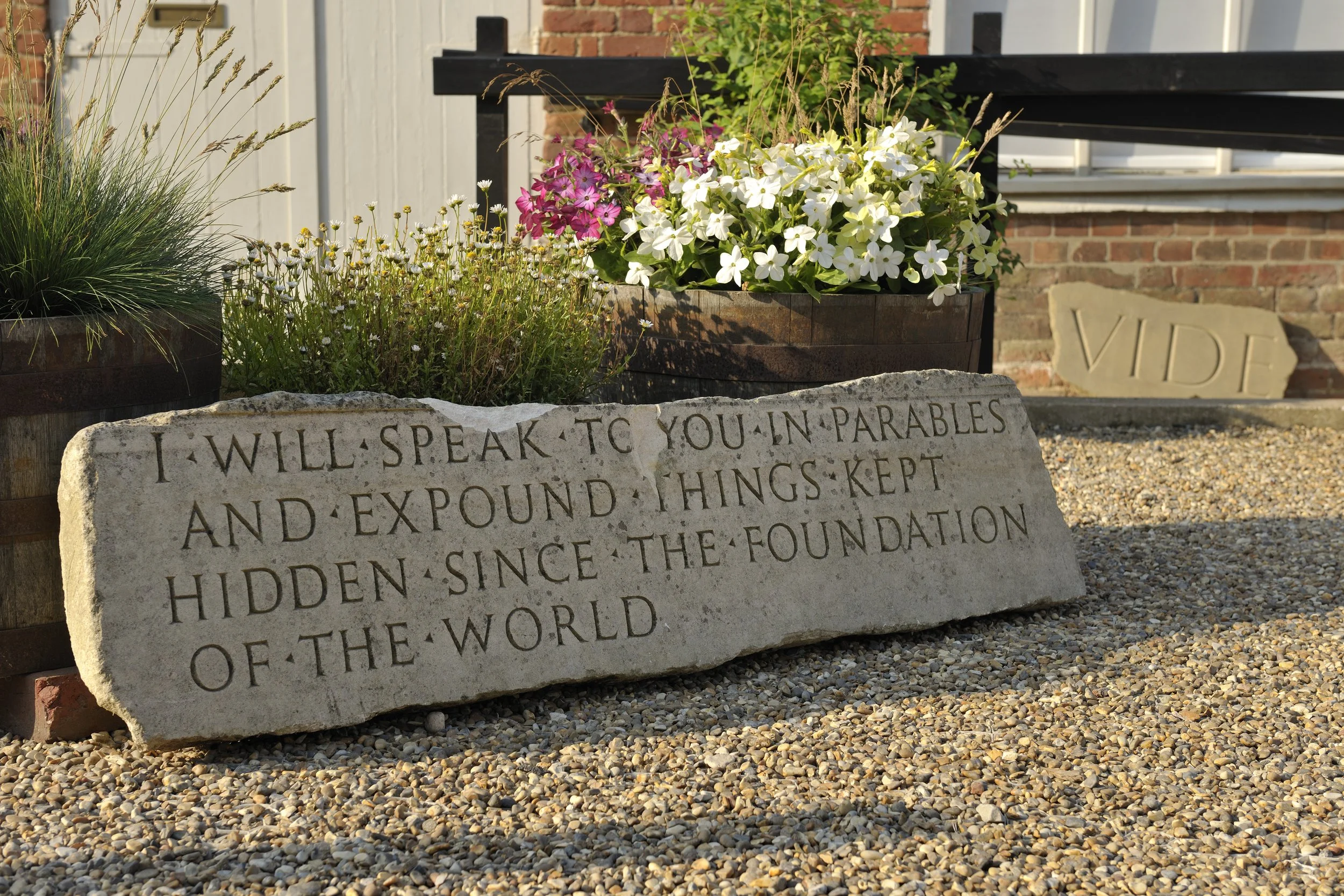 Stone memorial plaque with engraved text, surrounded by flower pots with pink and white flowers and greenery, placed on gravel ground outside a building with brick wall and white window/door frame.