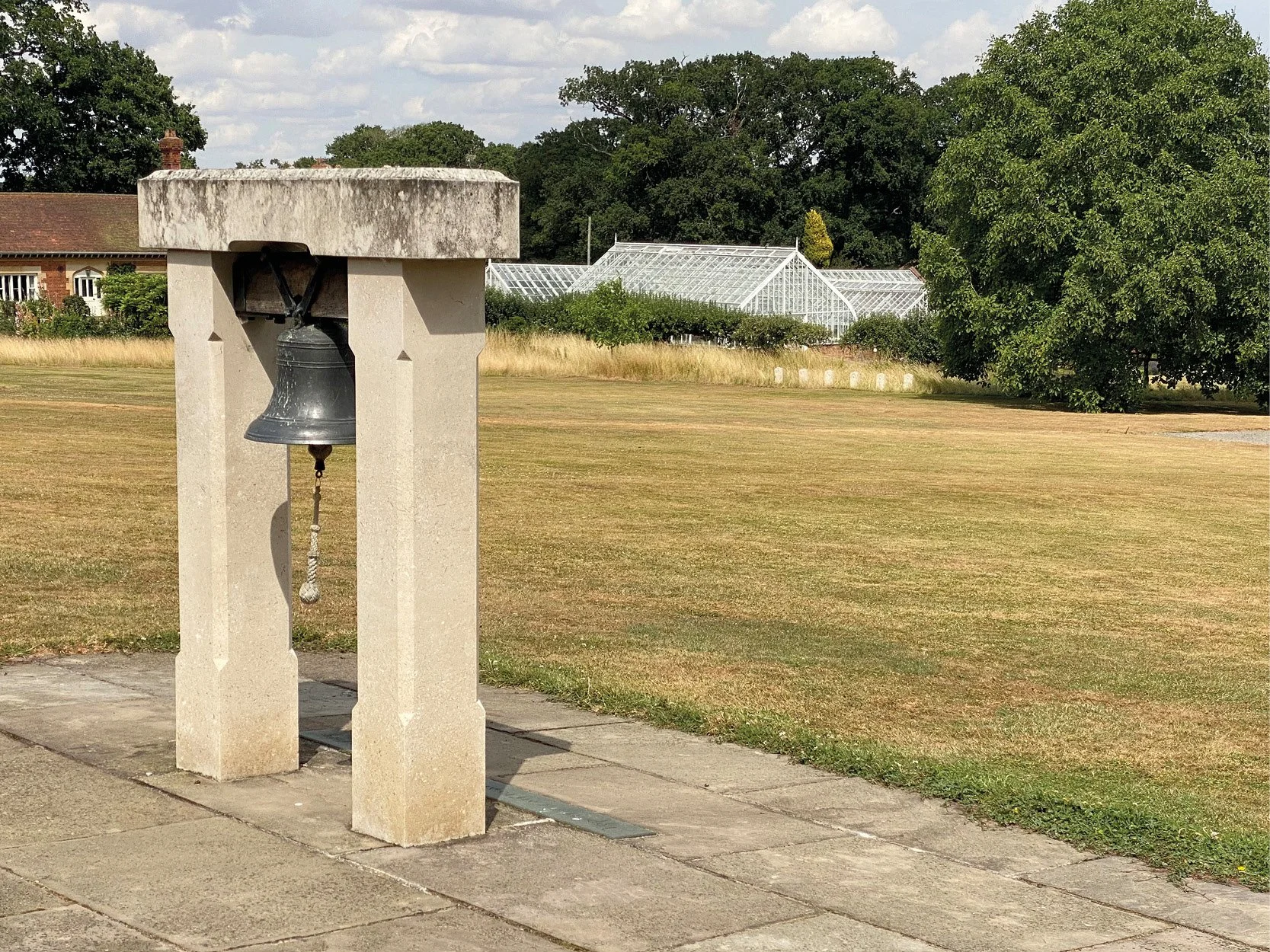 A black bell hanging under a concrete two-stand structure on a paved walkway. In the background, an open field with some trees, a greenhouse, and a partly cloudy sky.
