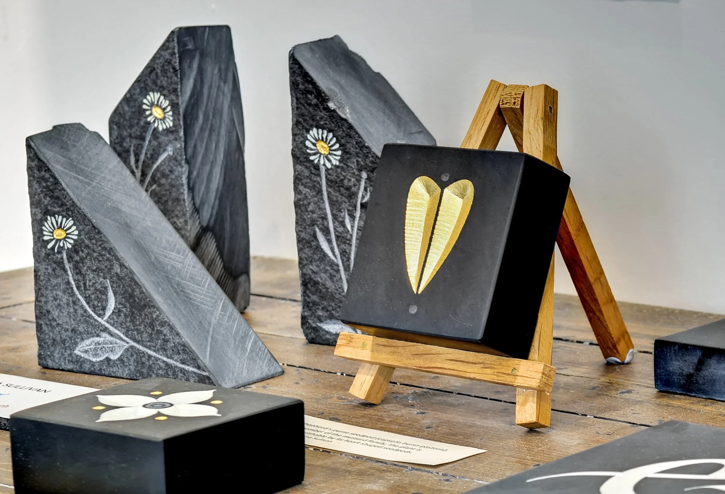 Several painted stone pieces featuring floral designs, displayed on a wooden table with a wooden easel in the background.