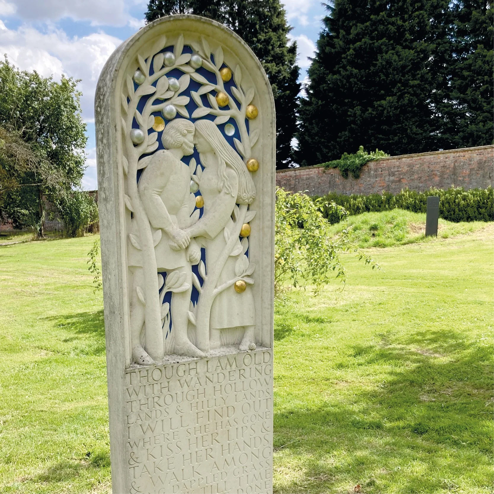 A marble memorial plaque with a carved relief of a couple kissing under a tree, surrounded by colorful spherical decorations, outdoors with green grass, trees, a brick wall, and cloudy sky in the background.
