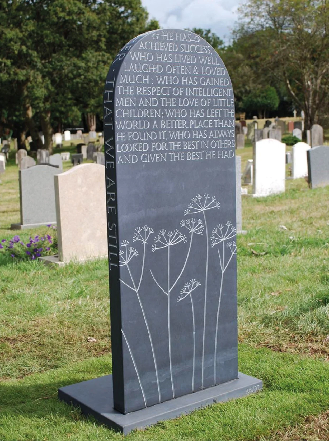 A black gravestone with an inscription and decorative flower illustrations in a cemetery with other headstones and trees in the background.