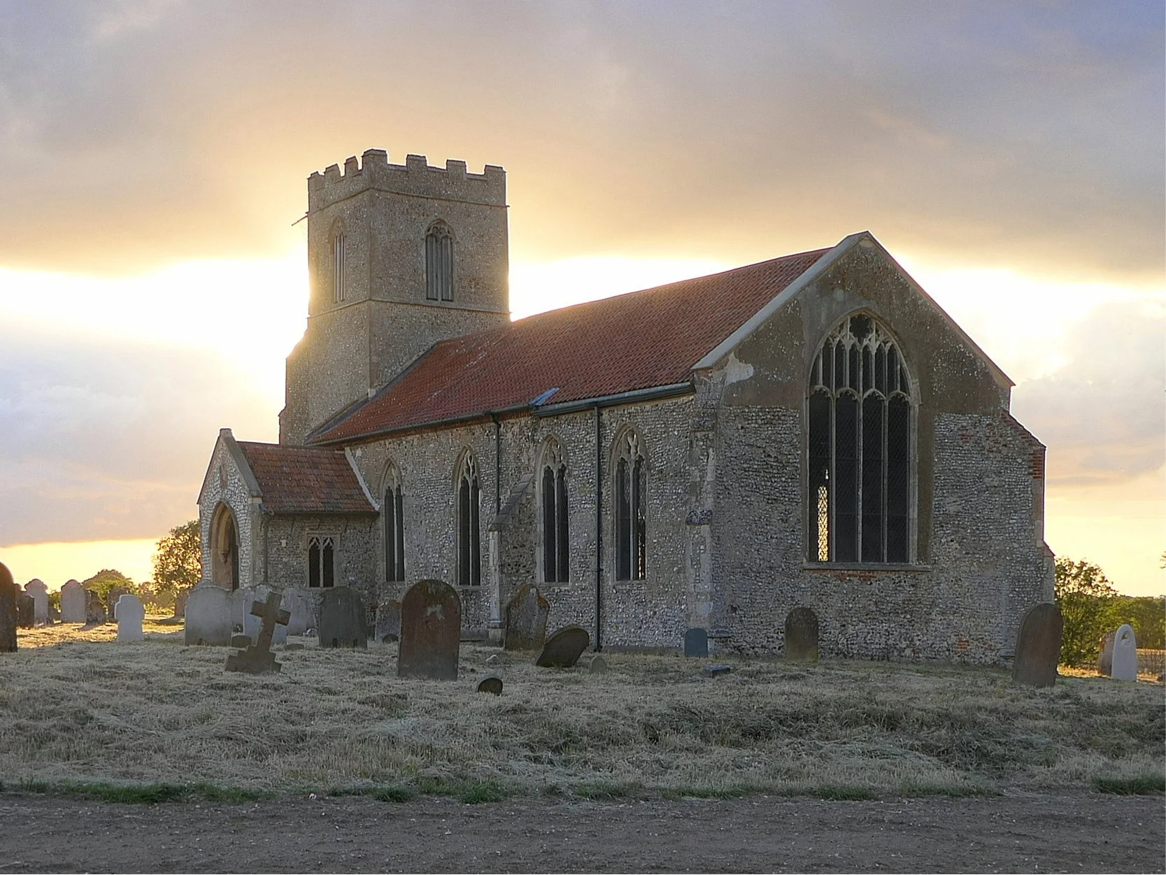 An old stone church with a graveyard in front, set against a sunset sky.