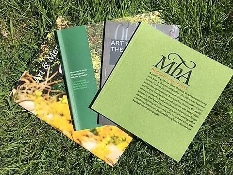 Four books or booklets lying on grass, one with a green cover and the others with art or photography covers.