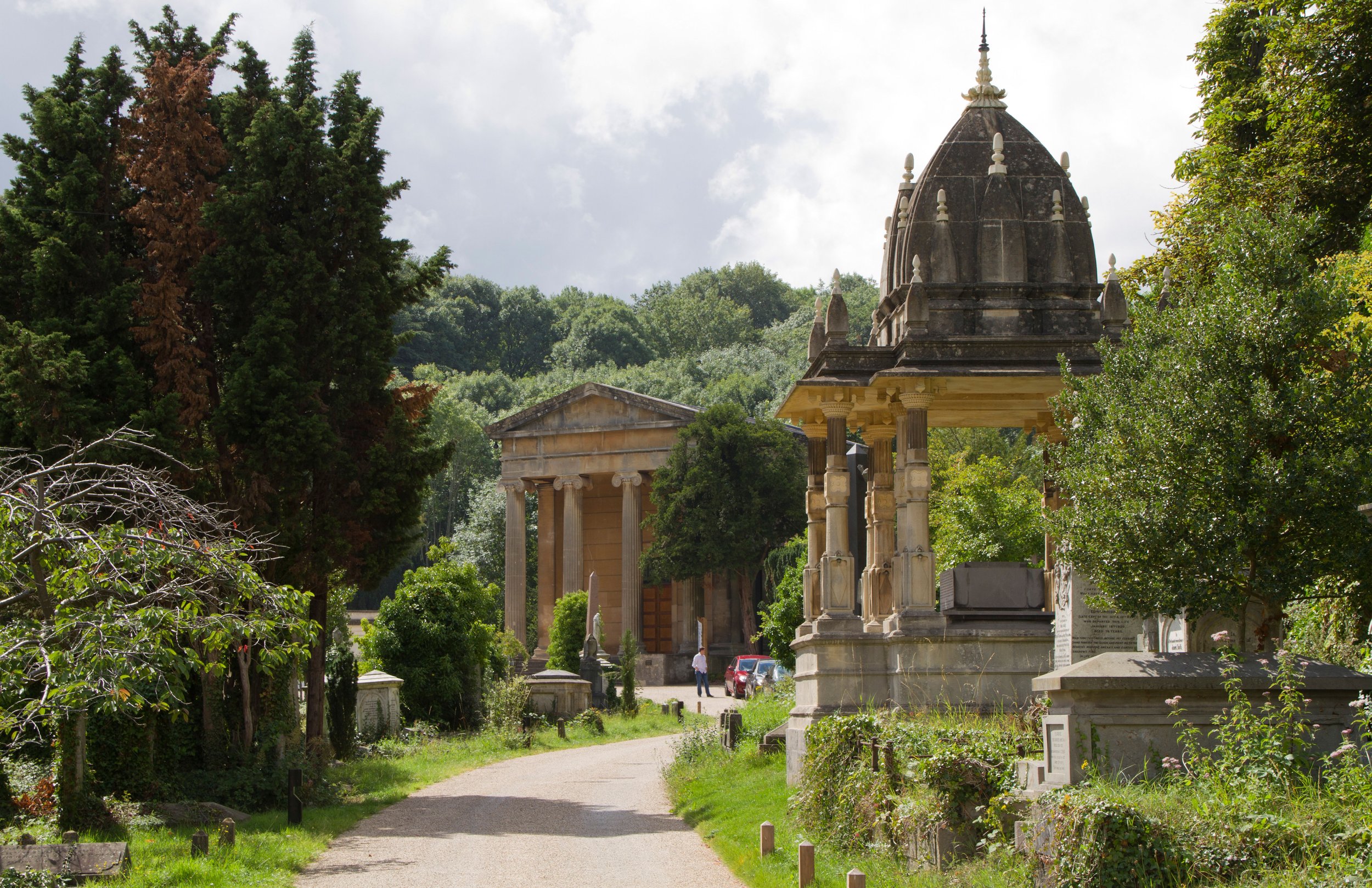 Historical gravesite surrounded by lush green trees, with a pathway leading through it. A person is seen walking among the stone structures, and an ornate, domed monument is prominent on the right side of the scene.