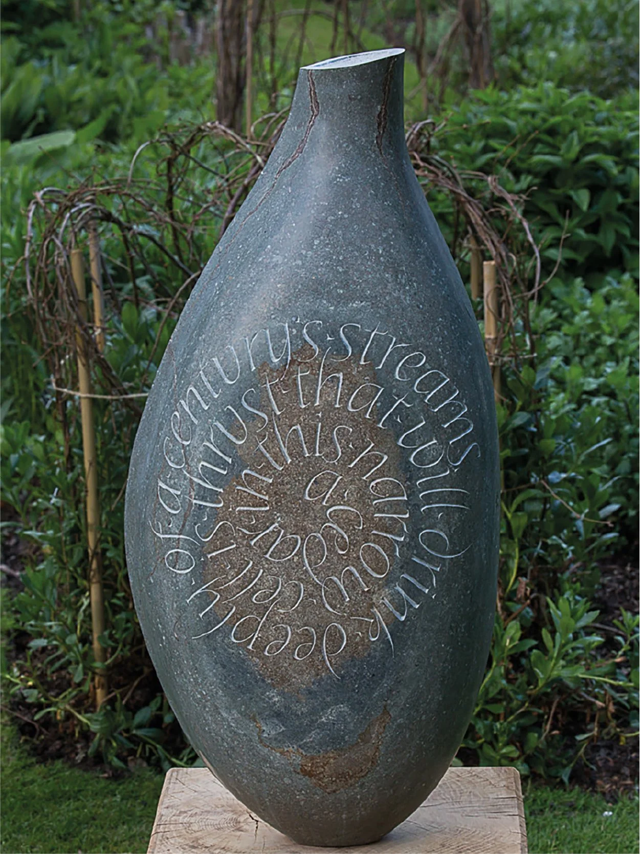 A large, dark, elongated ceramic sculpture with spiral engraved text in a garden setting with green foliage and vines in the background.