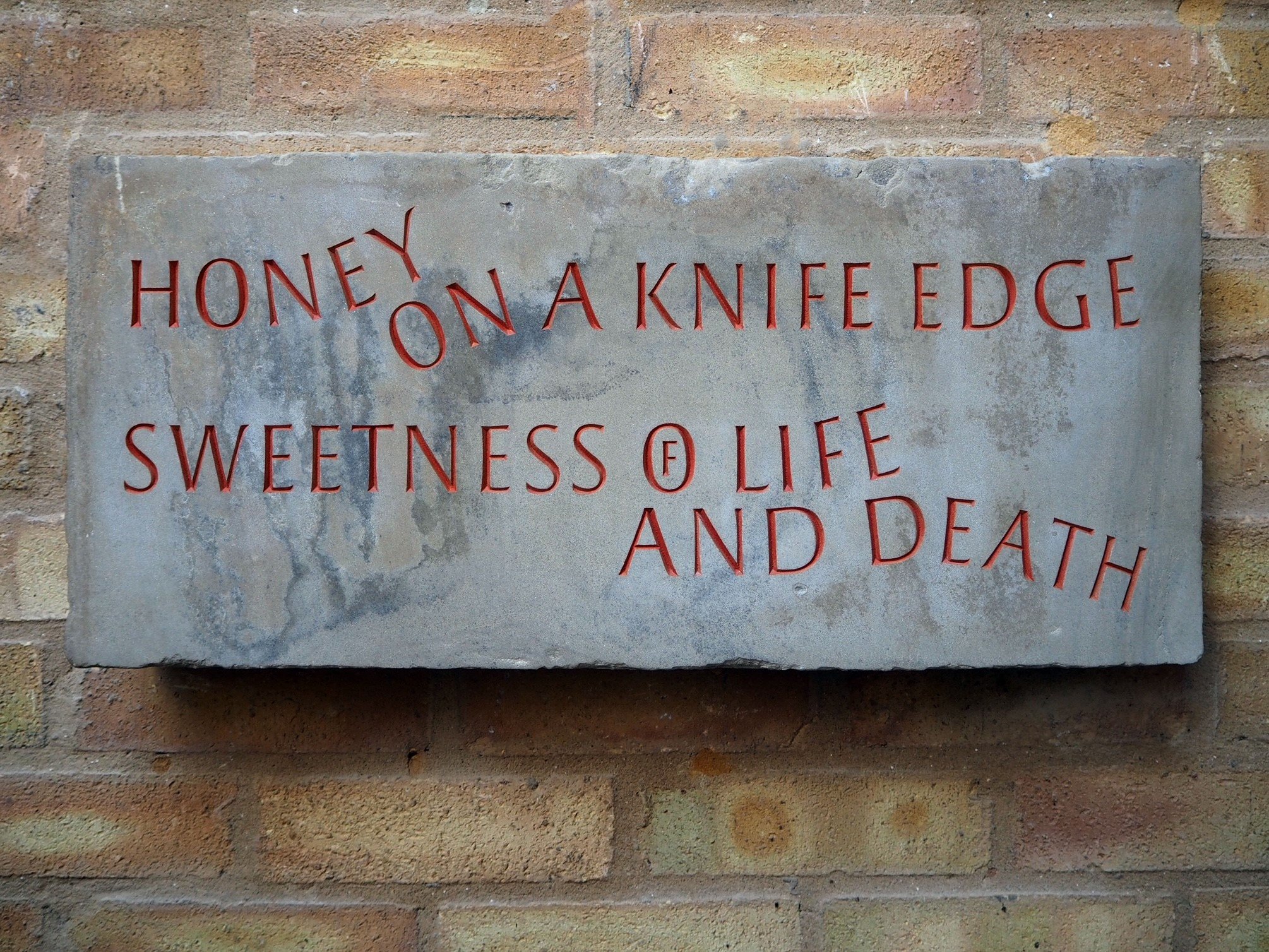 A rectangular wall plaque with red embossed text on a gray stone background, mounted on a brick wall. The text reads: 'HONEY ON A KNIFE EDGE SWEETNESS OF LIFE AND DEATH'.