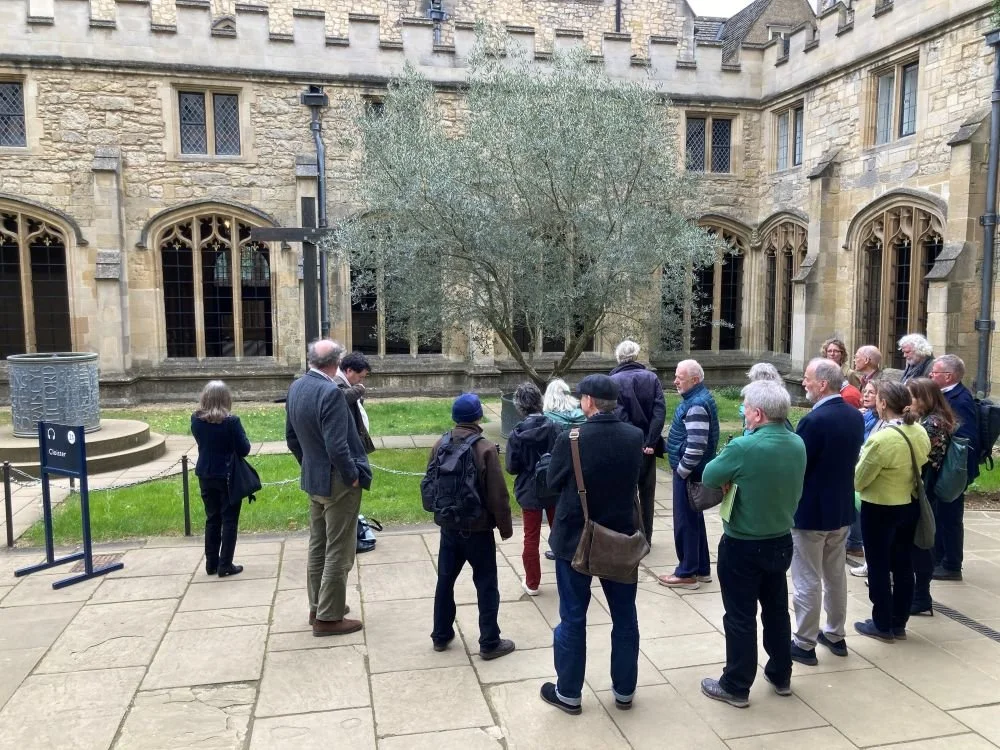 Group of people listening to a guide outside a historic stone building with tall windows, in a courtyard with a large tree and green grass.