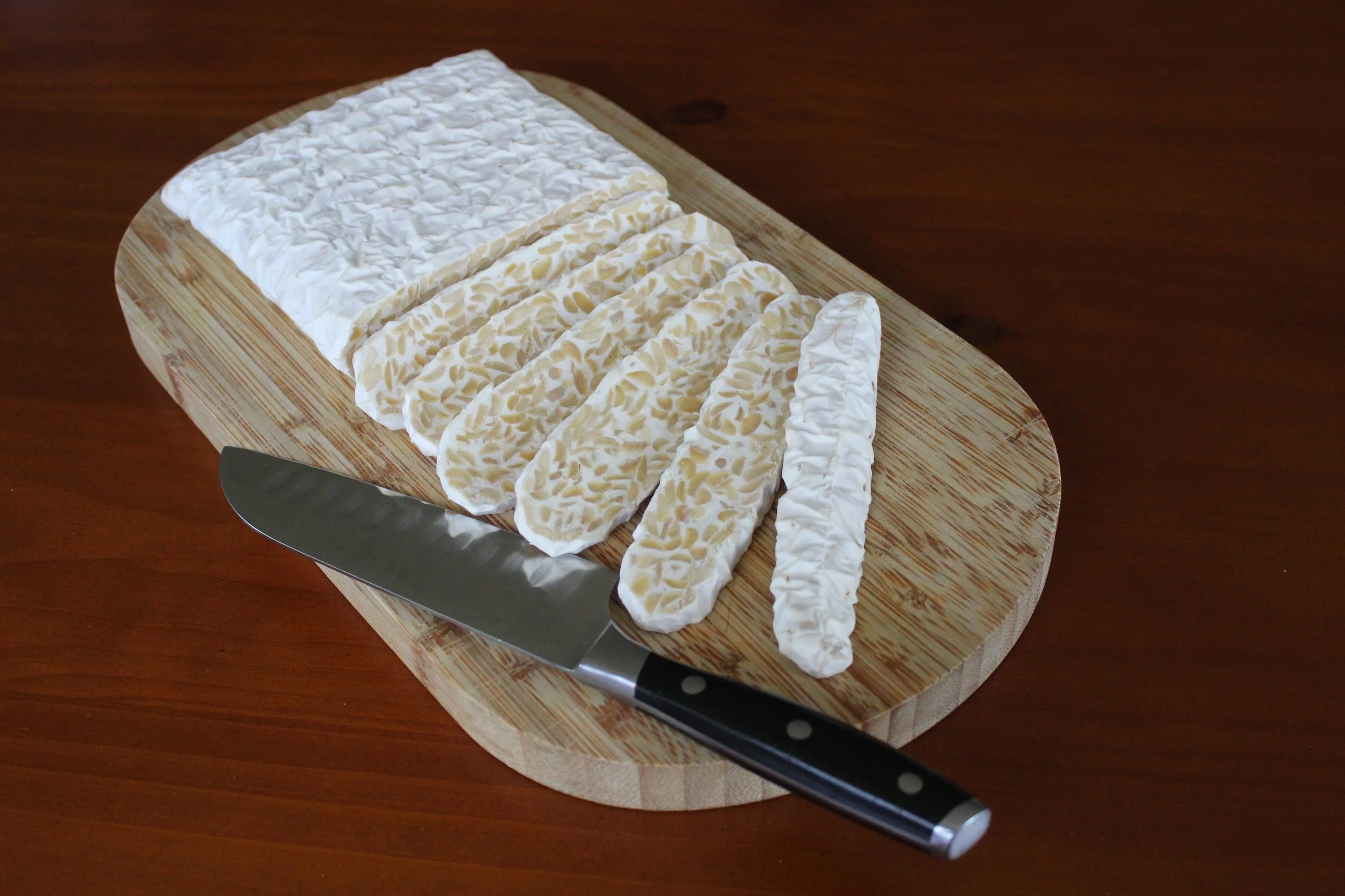 Blocks of sliced tempeh on a wooden cutting board with a black-handled knife resting on the board, on a wooden surface.
