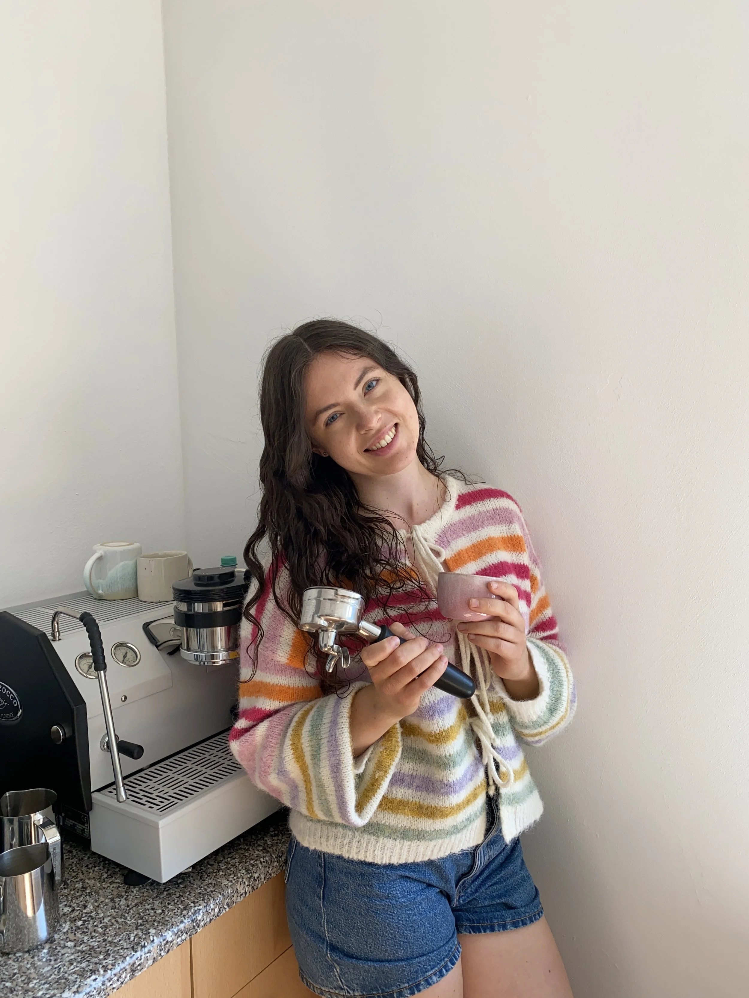 Eine junge Frau mit langen dunklen Haaren, die in einem farbenfrohen gestreiften Pullover und Shorts steht, eine Espressomaschine in der Hand und eine Tasse in der anderen, vor einer Küchenarbeitsplatte.
