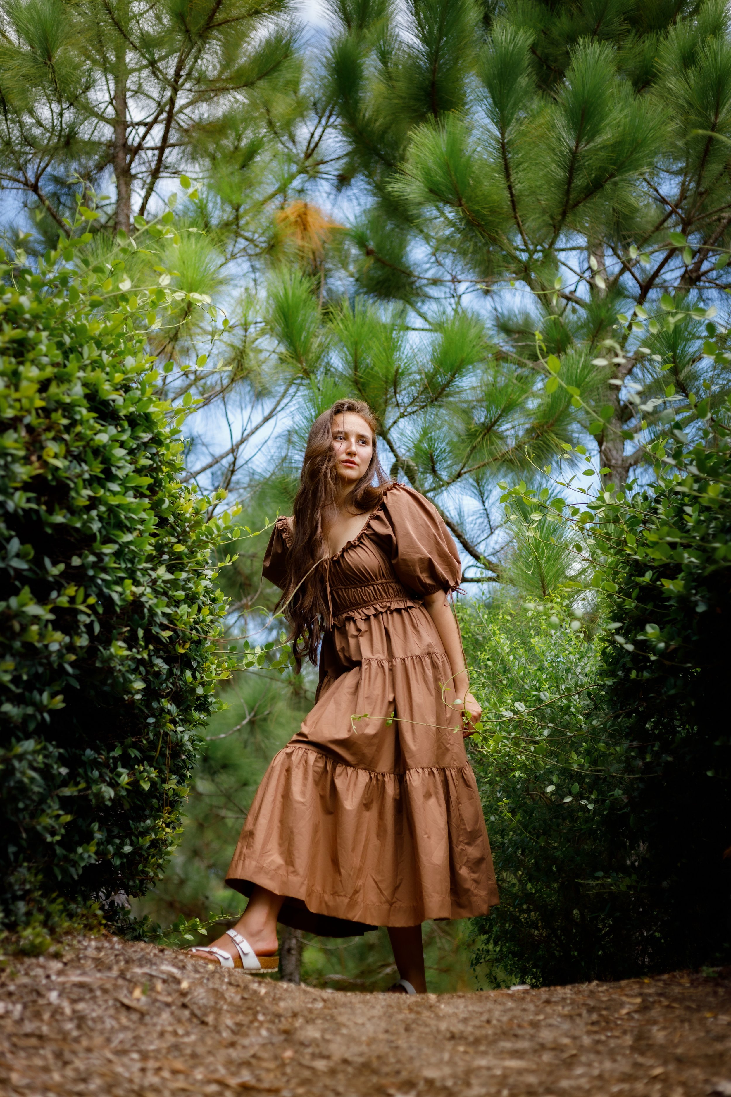 A woman in a brown dress standing on a dirt path surrounded by green bushes and tall pine trees.