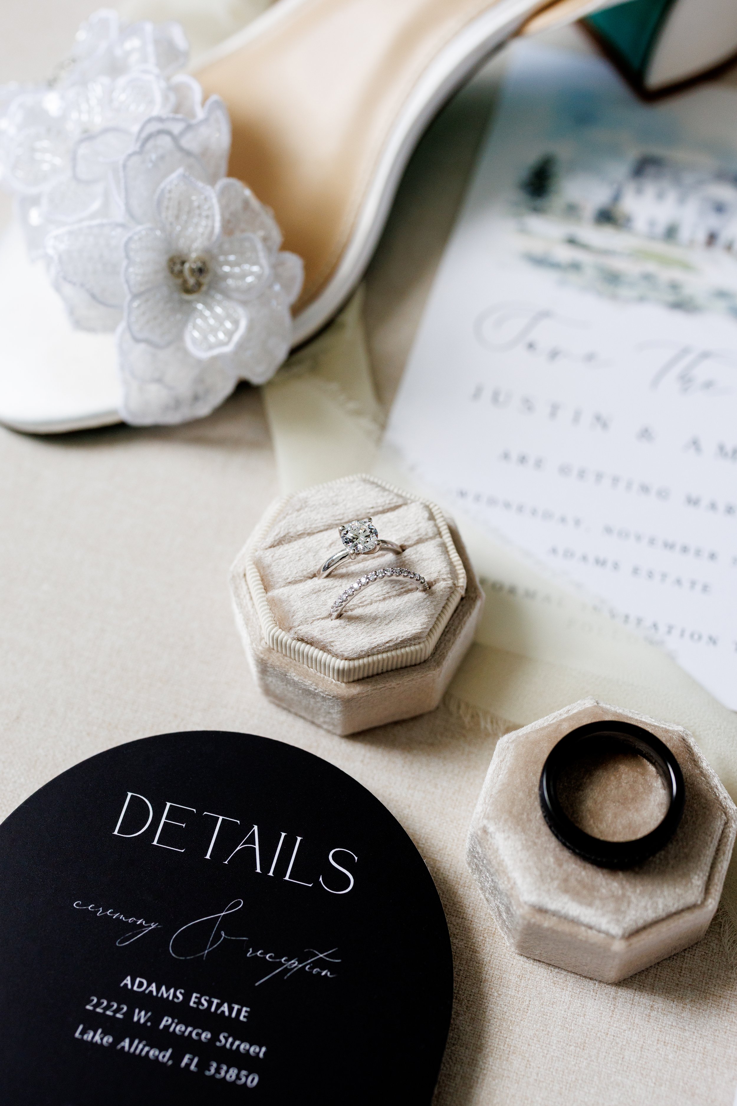 Close-up of a wedding planning scene with wedding rings in a plush ring box, a black wedding band, a black details card, and decorative flowers, with part of a wedding invitation visible in the background.