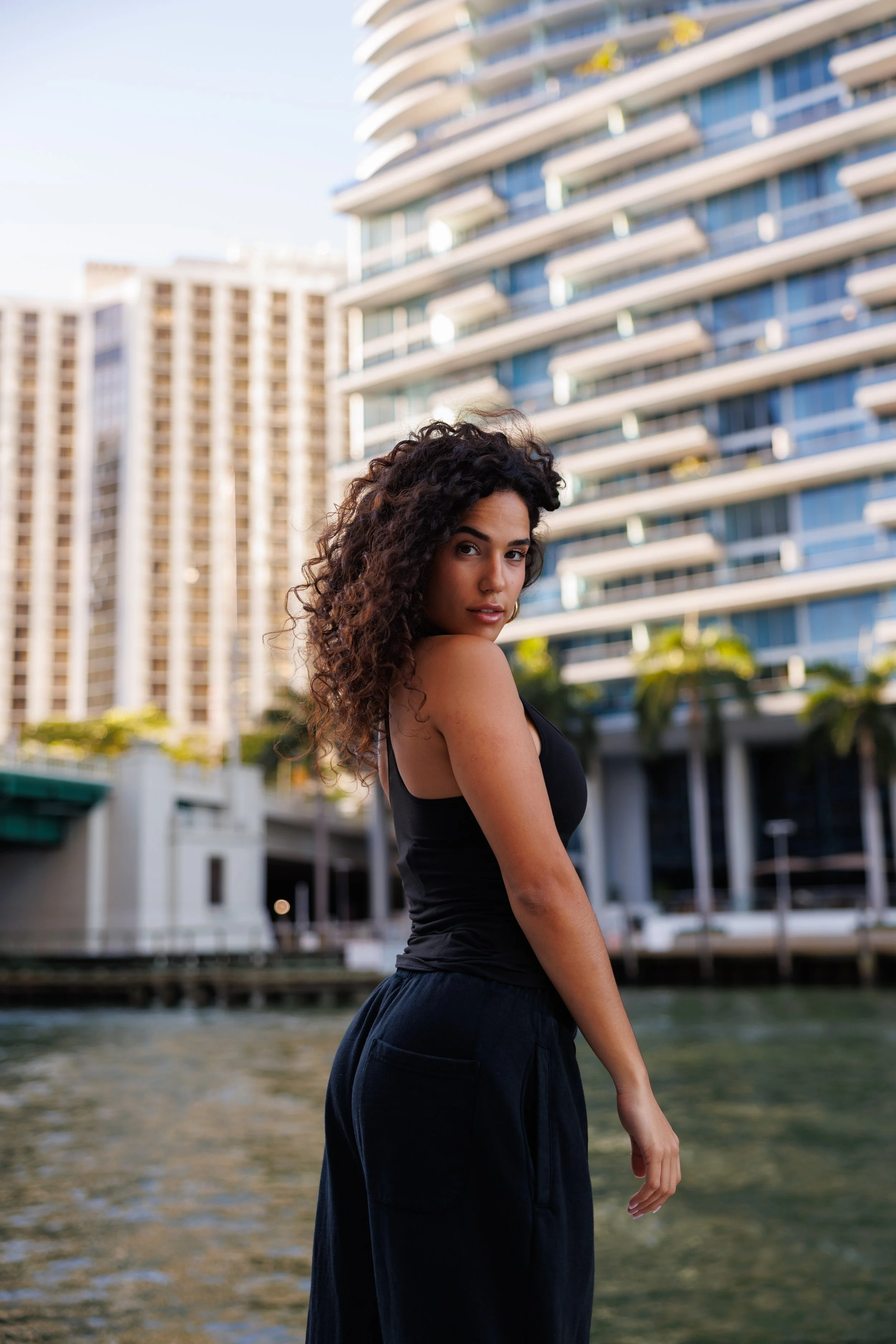 A woman with curly dark hair stands near a body of water with modern high-rise buildings in the background.