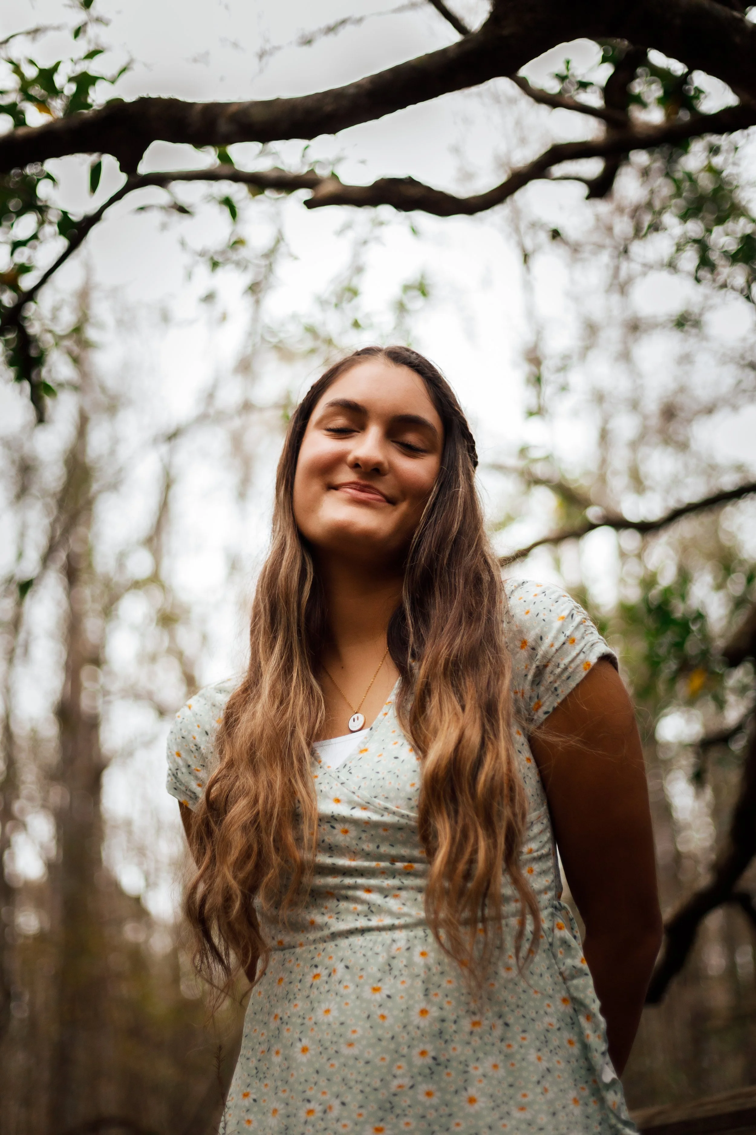 A young woman with long wavy hair and a content smile, standing outdoors in a wooded area, wearing a light-colored dress with small floral patterns.