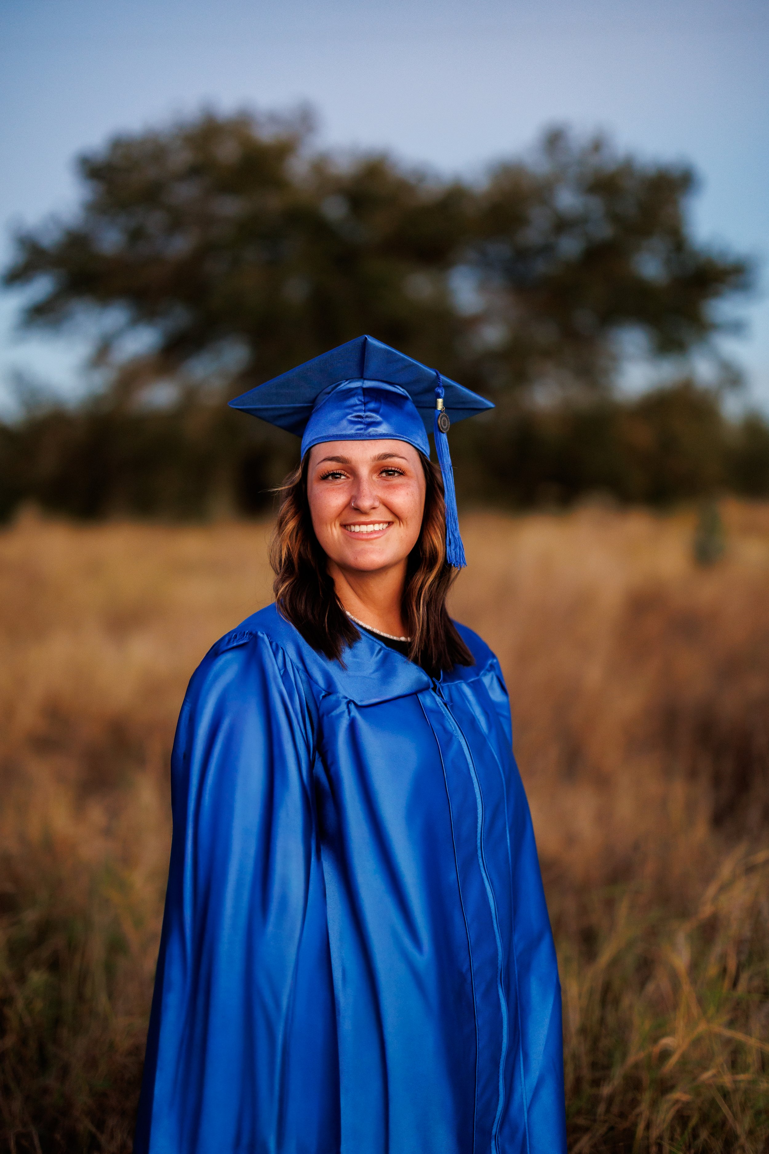 A young woman in a bright blue graduation gown and cap, smiling outdoors in a field with a large tree in the background during the evening.
