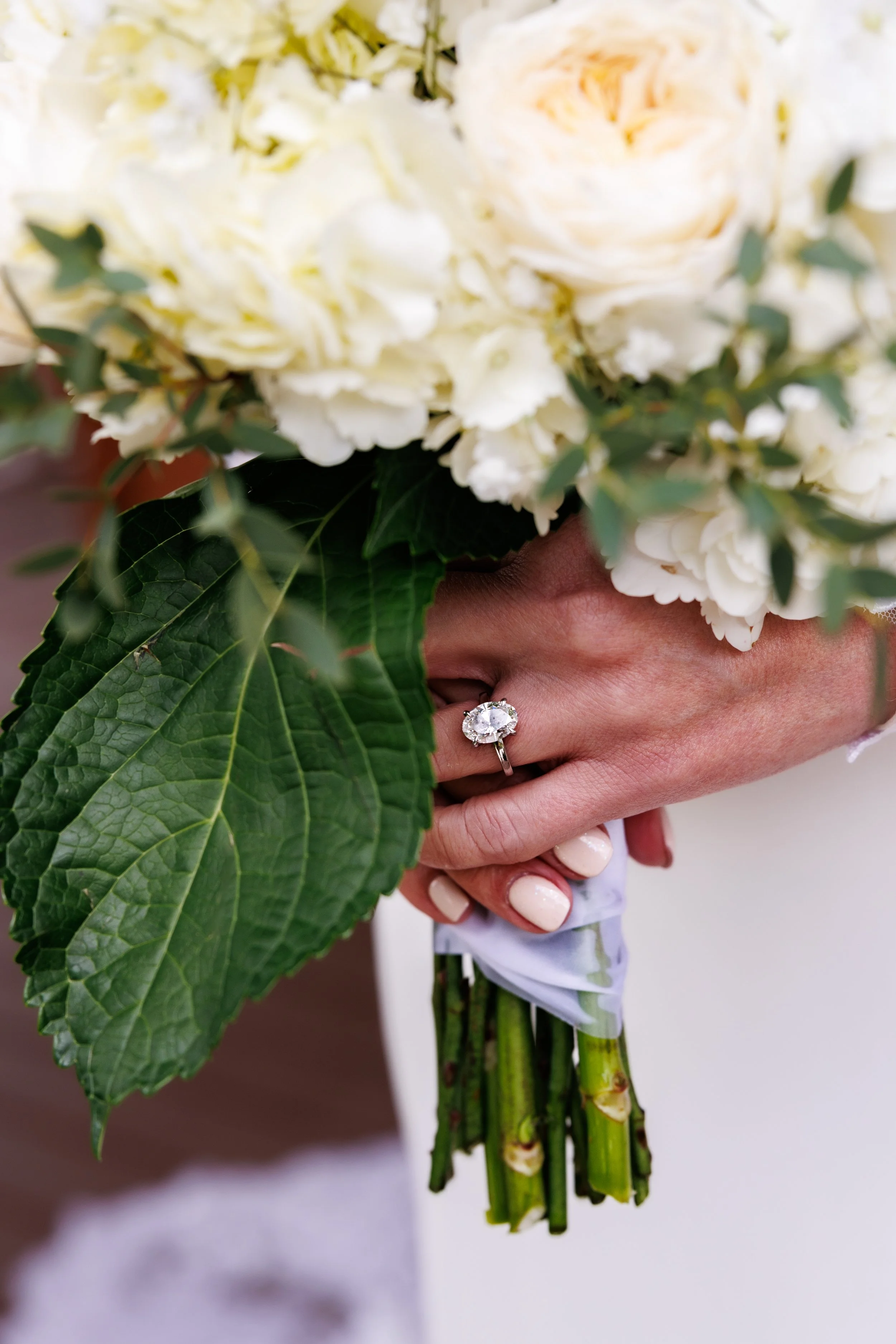 Close-up of a person holding a bouquet of white flowers, including hydrangeas and roses, with a large green leaf covering part of their hand, and an engagement ring on their finger.