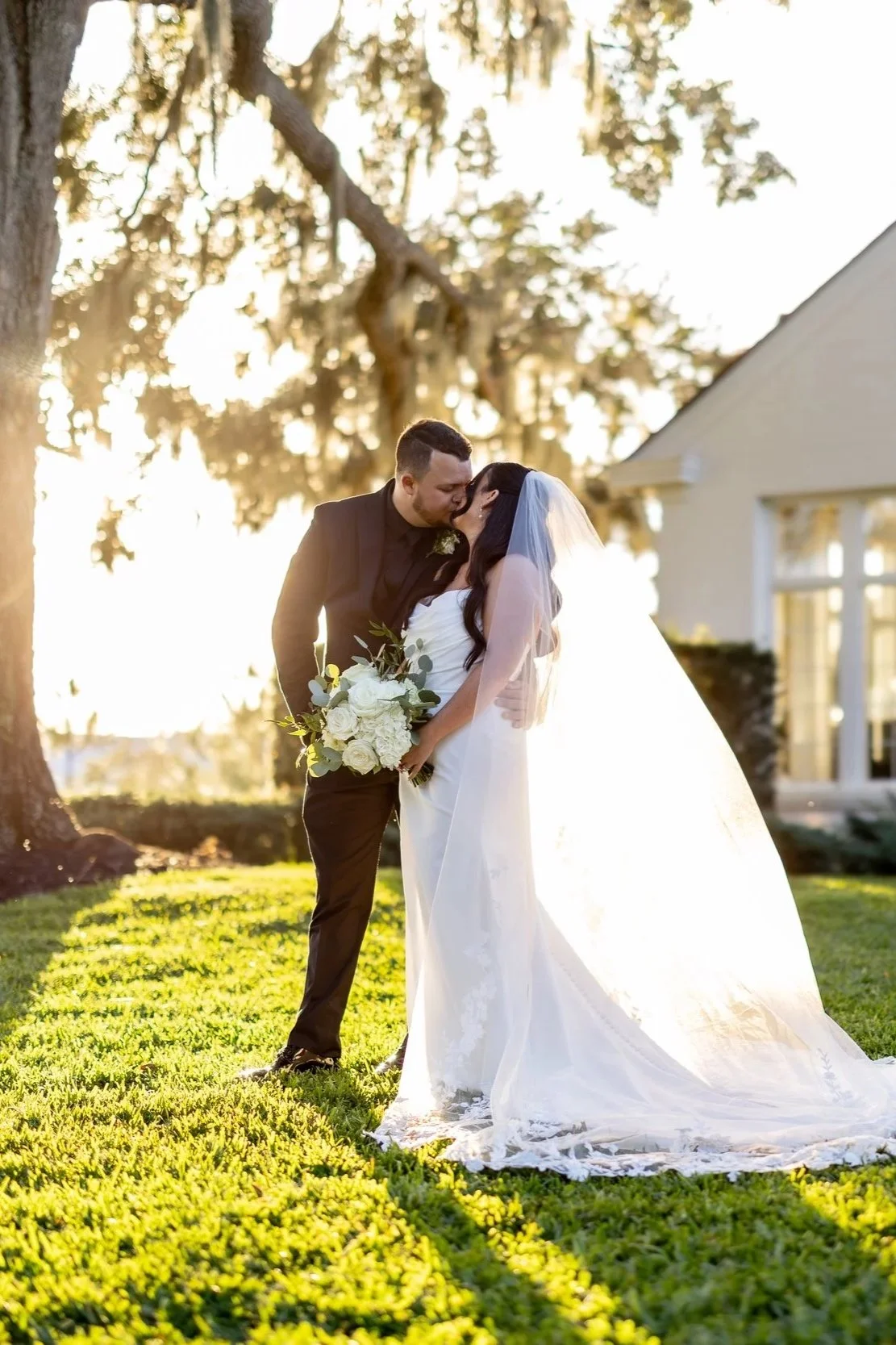 A bride and groom sharing a kiss outdoors during sunset, with sunlight filtering through trees, the bride in a white wedding gown holding a bouquet of white roses, and the groom in a black suit.