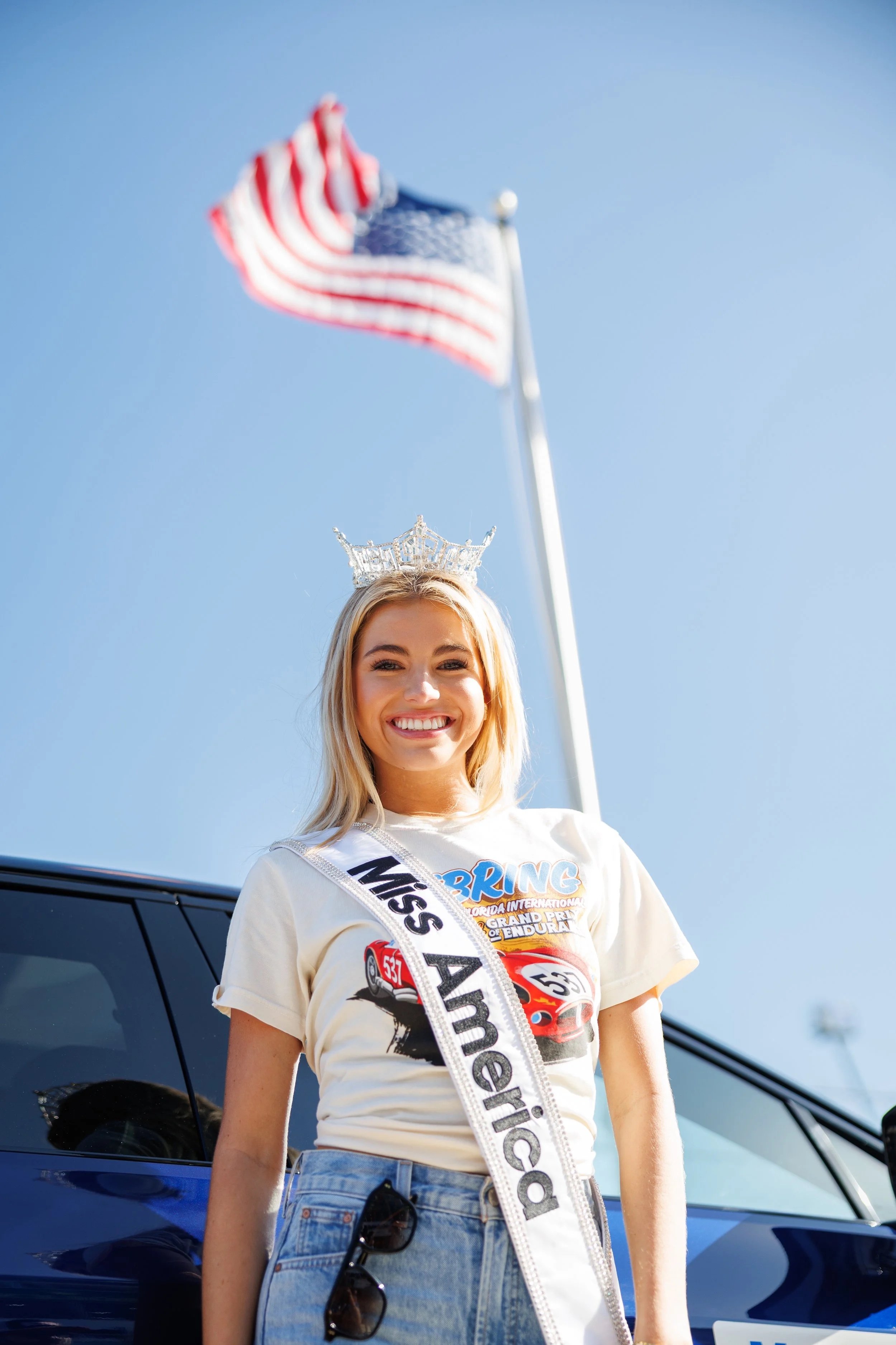 Young woman smiling, wearing a Miss America sash and a crown, standing outdoors with an American flag in the background.