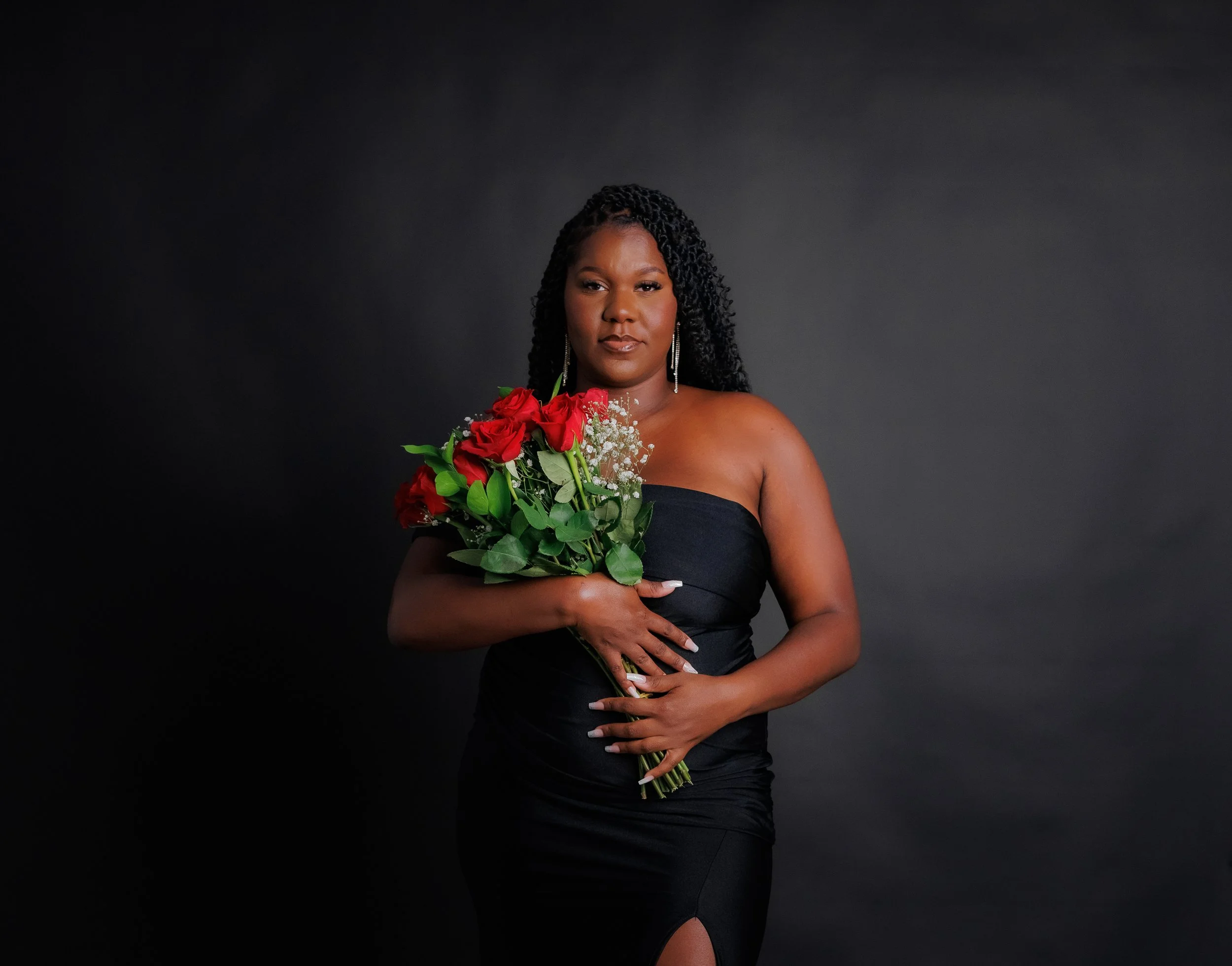 Woman in a strapless black dress holding a bouquet of red roses and white baby's breath flowers against a dark background.