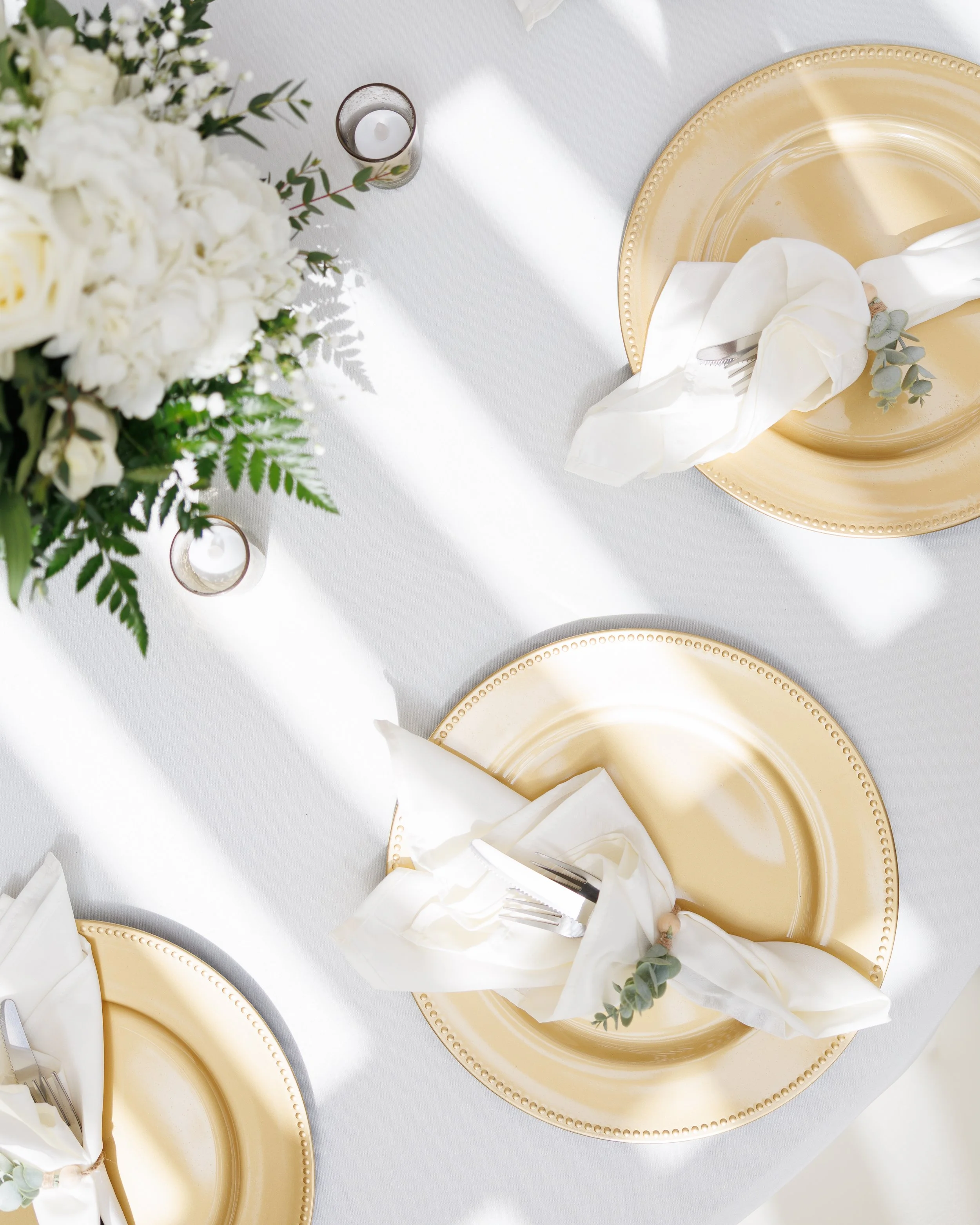 Table setting with gold plates, white napkins, forks, and a floral centerpiece with white flowers and greenery, illuminated by natural light.