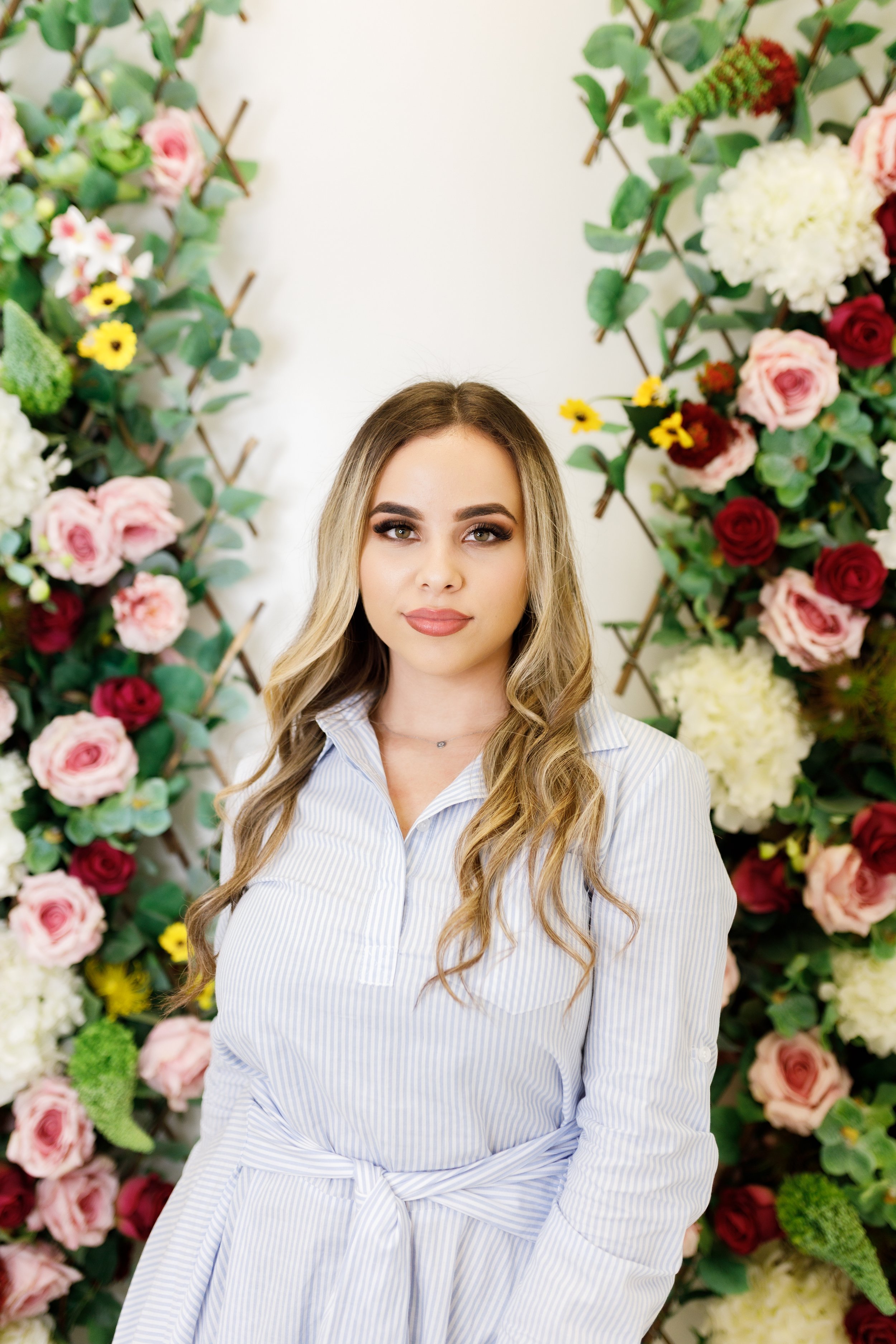 A young woman with long wavy hair and makeup, wearing a light blue striped dress, standing in front of a floral backdrop with pink, red, white, and yellow flowers and green leaves.