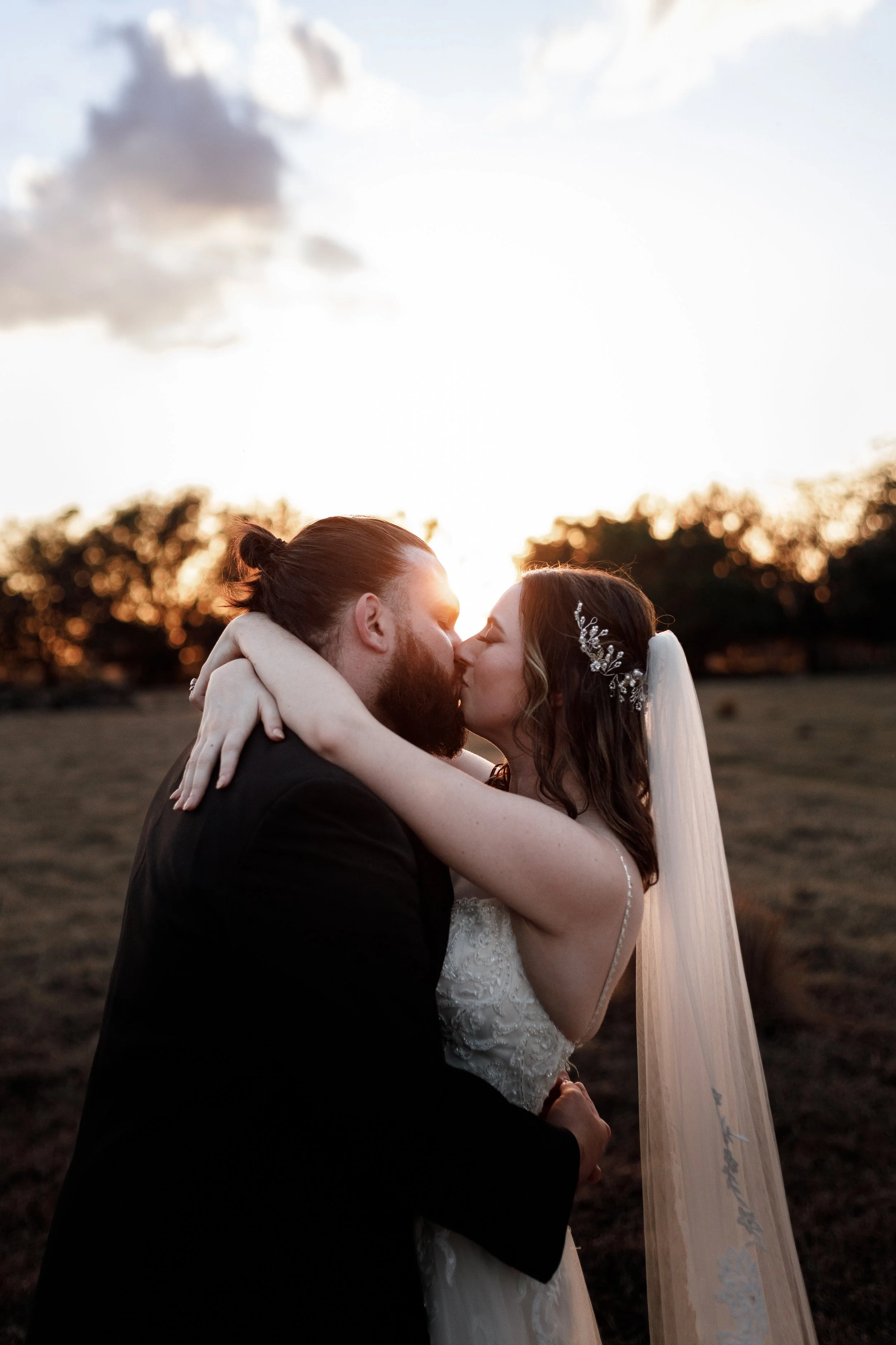 A bride and groom sharing a kiss outdoors at sunset, with trees in the background.