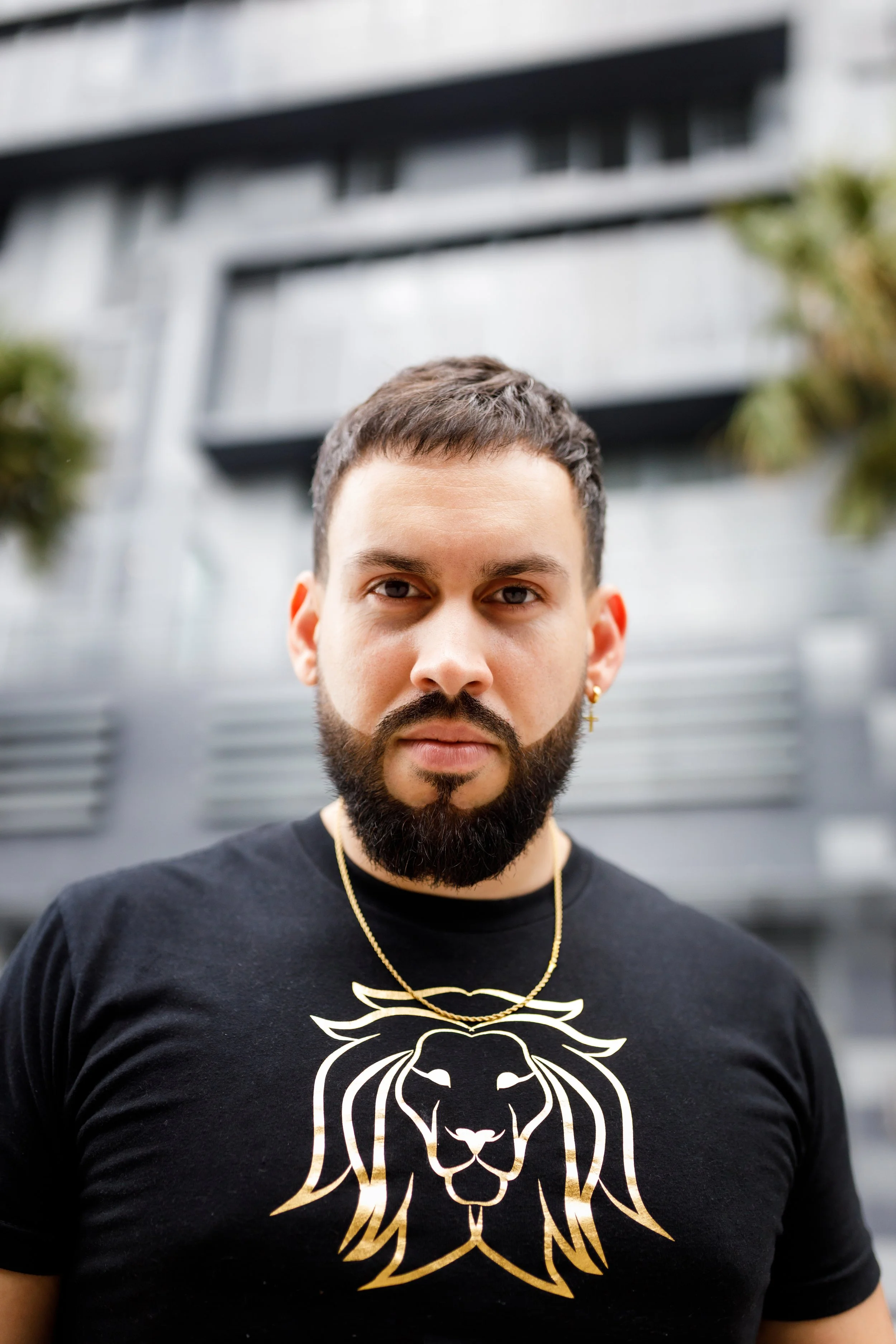 Close-up of a man with a beard, wearing a black t-shirt with a gold lion print and gold jewelry, standing outdoors in front of a modern building.