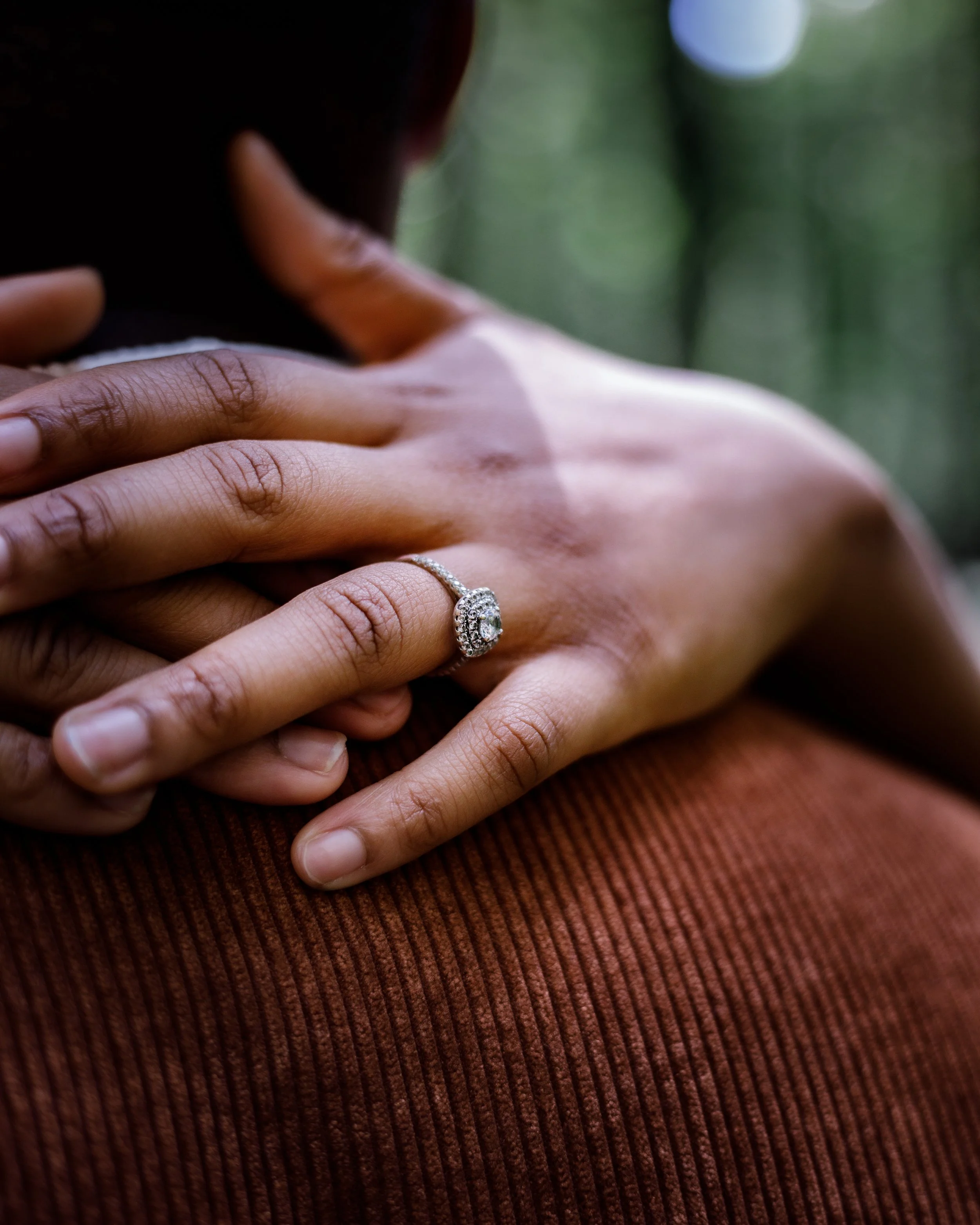 A close-up of a person's hand showing an engagement ring, resting on another person's shoulder in an outdoor setting.