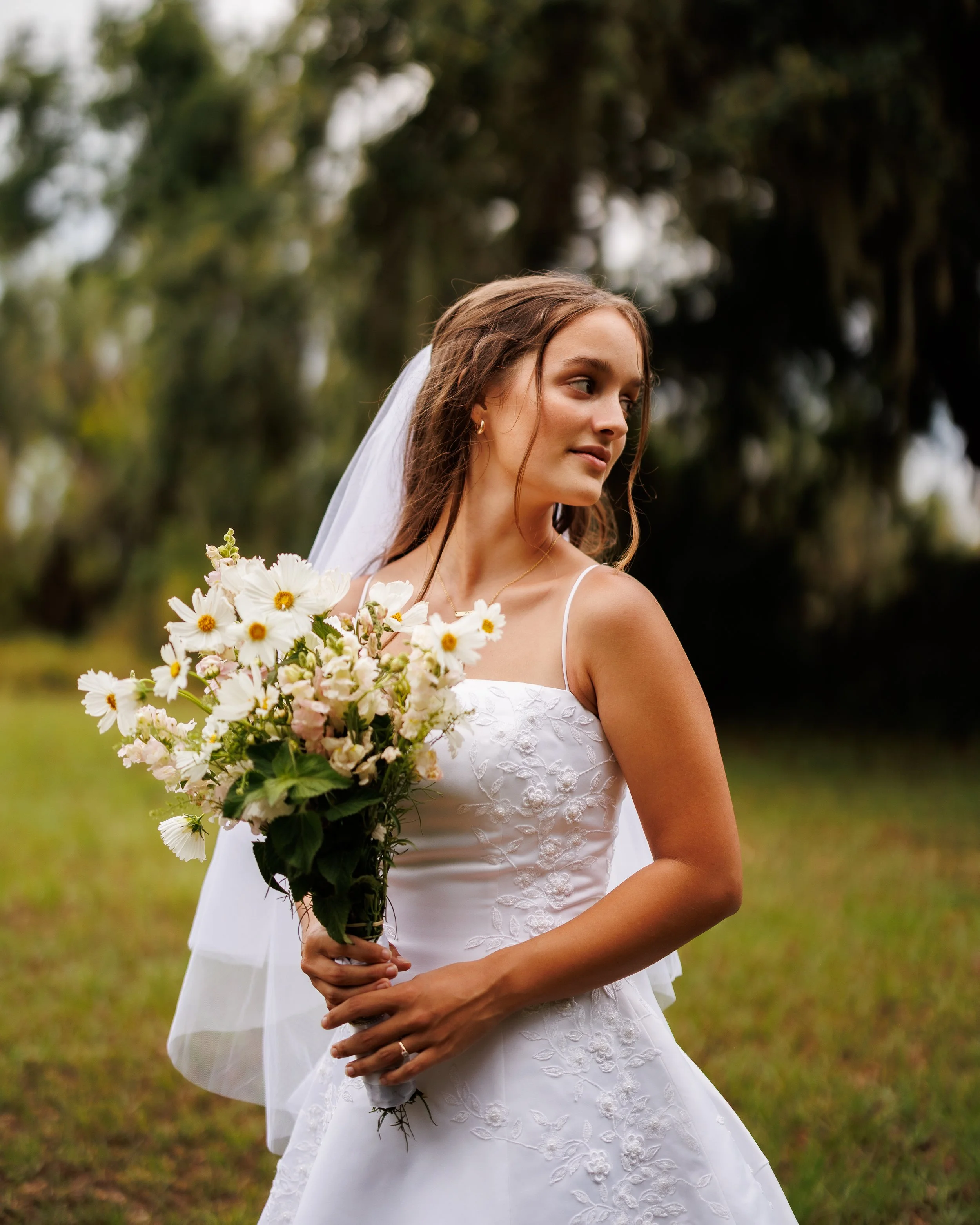 A bride in a white wedding gown holding a bouquet of white and pink flowers outdoors with trees in the background.