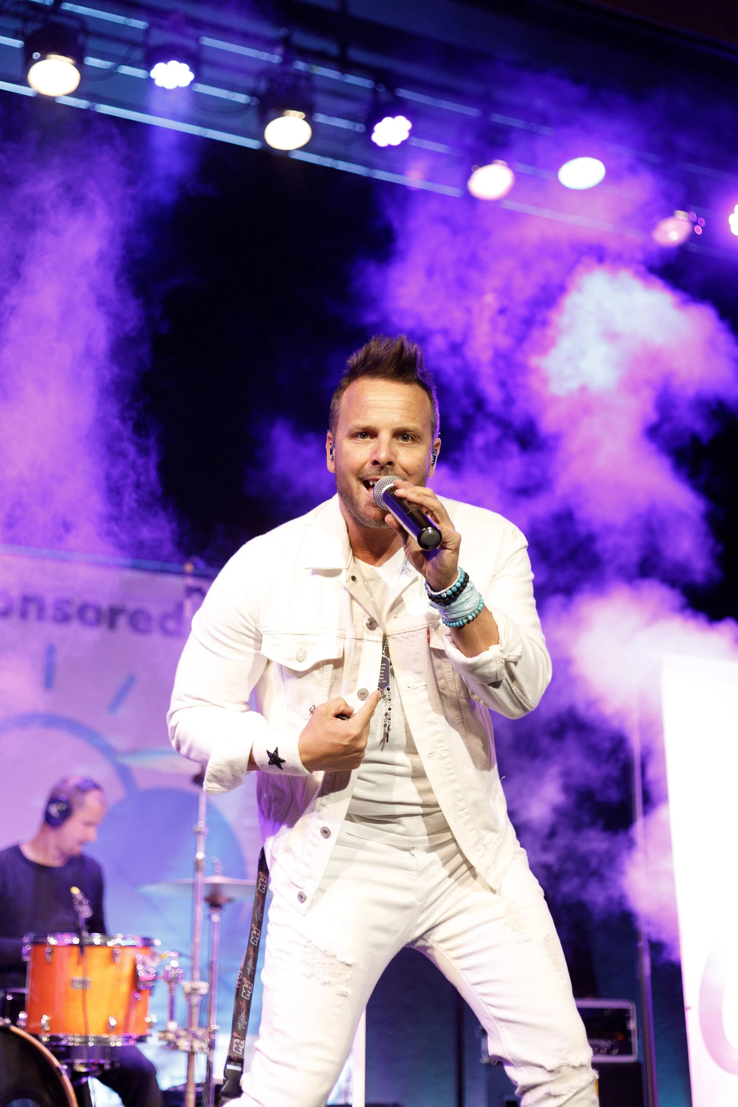 Male singer in white denim outfit with bracelets, performing on stage with purple lighting and smoke effects, and a drummer in the background.