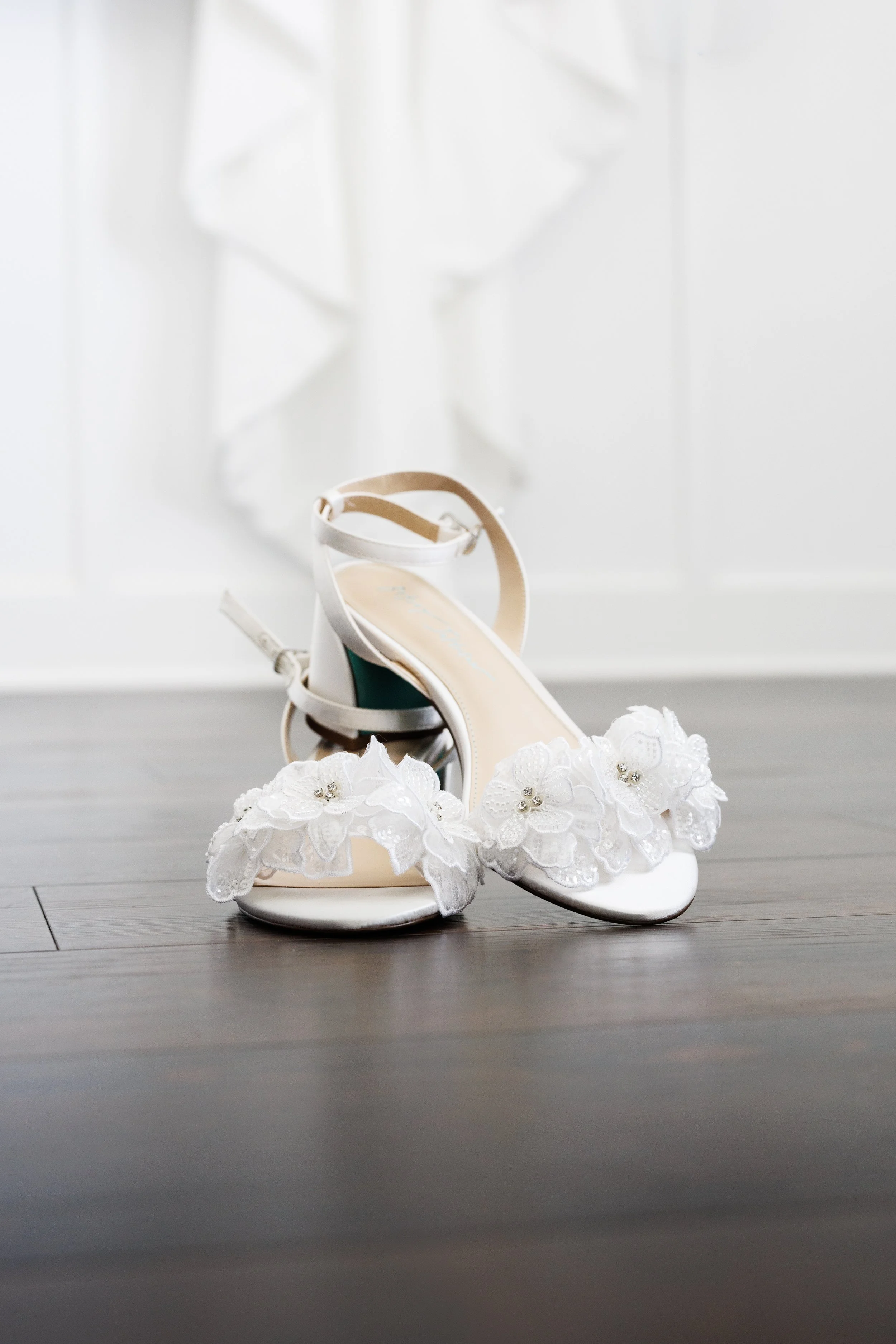 A pair of white wedding shoes with floral lace details on the front, placed on a dark wooden floor with a white background.