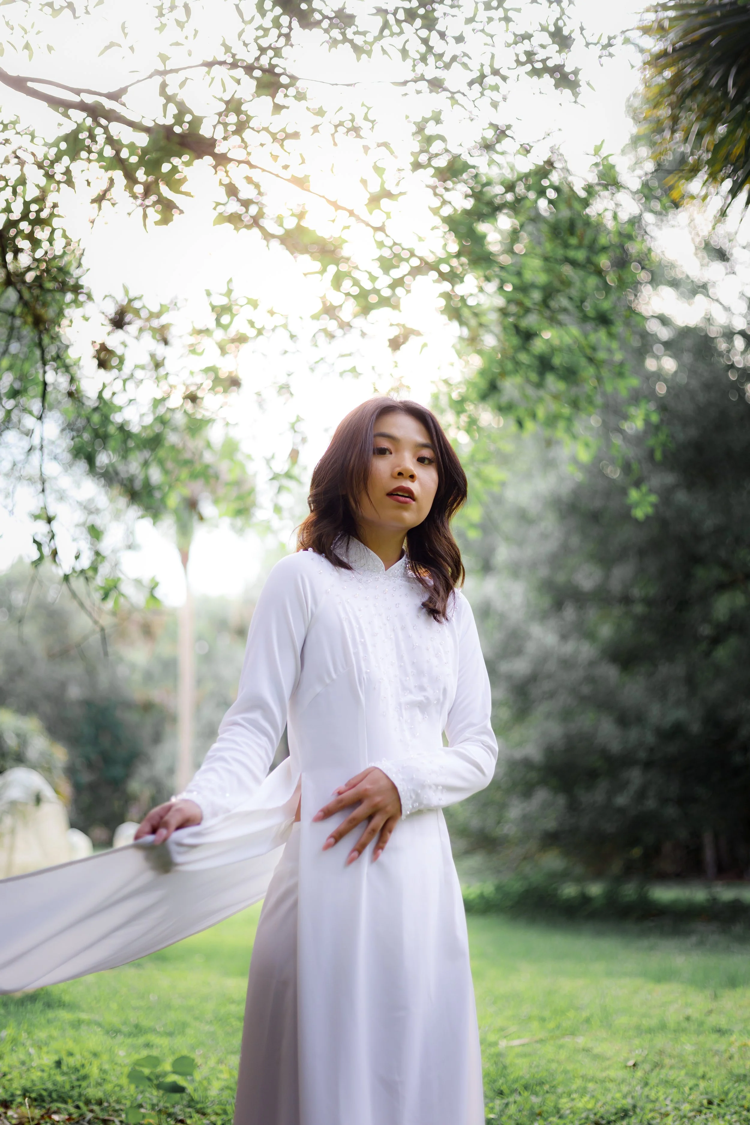 A young woman in a white dress standing outdoors in a lush green park, with sunlight filtering through the trees.