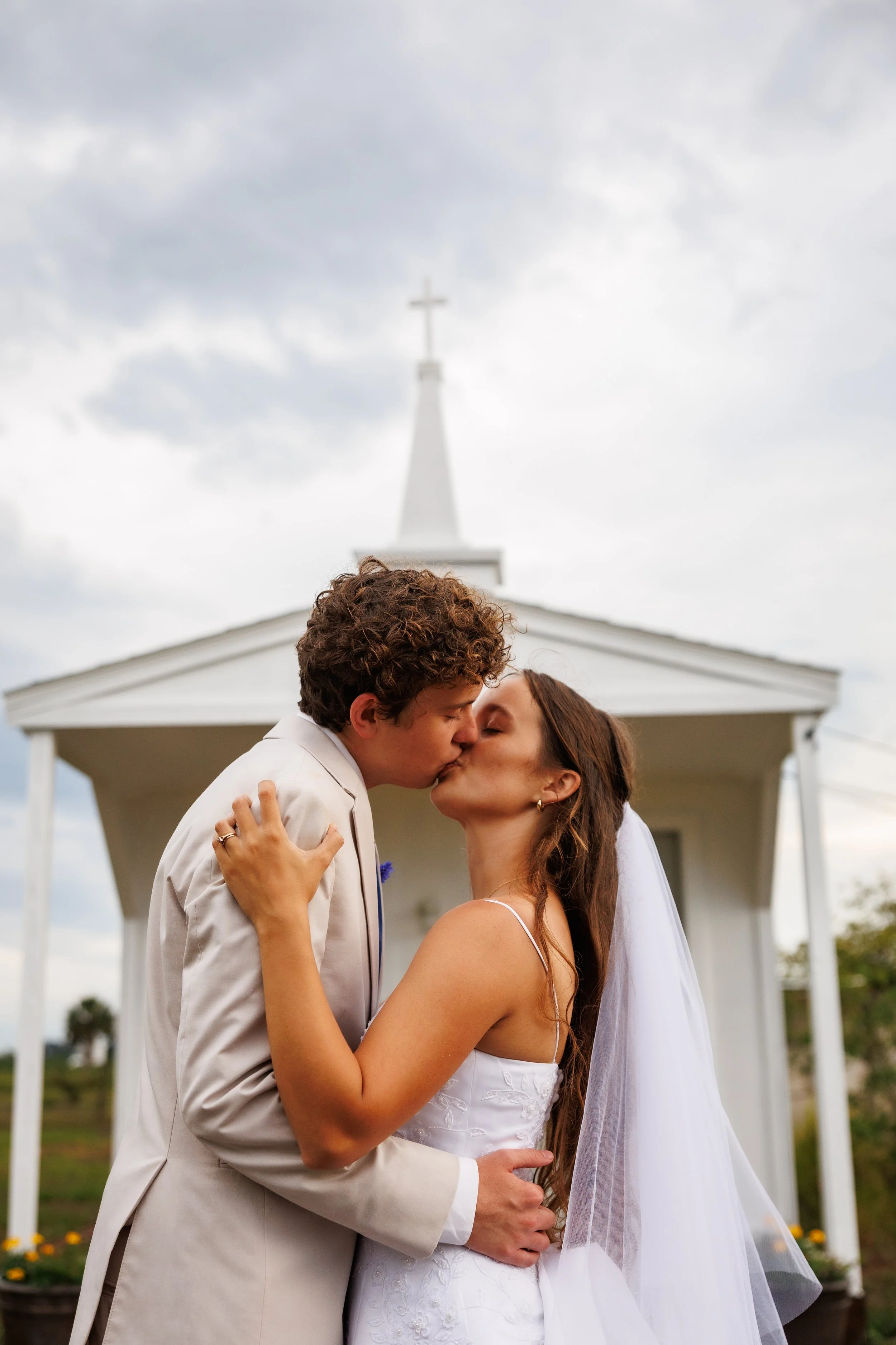 A bride and groom kiss in front of a white chapel during their wedding ceremony.