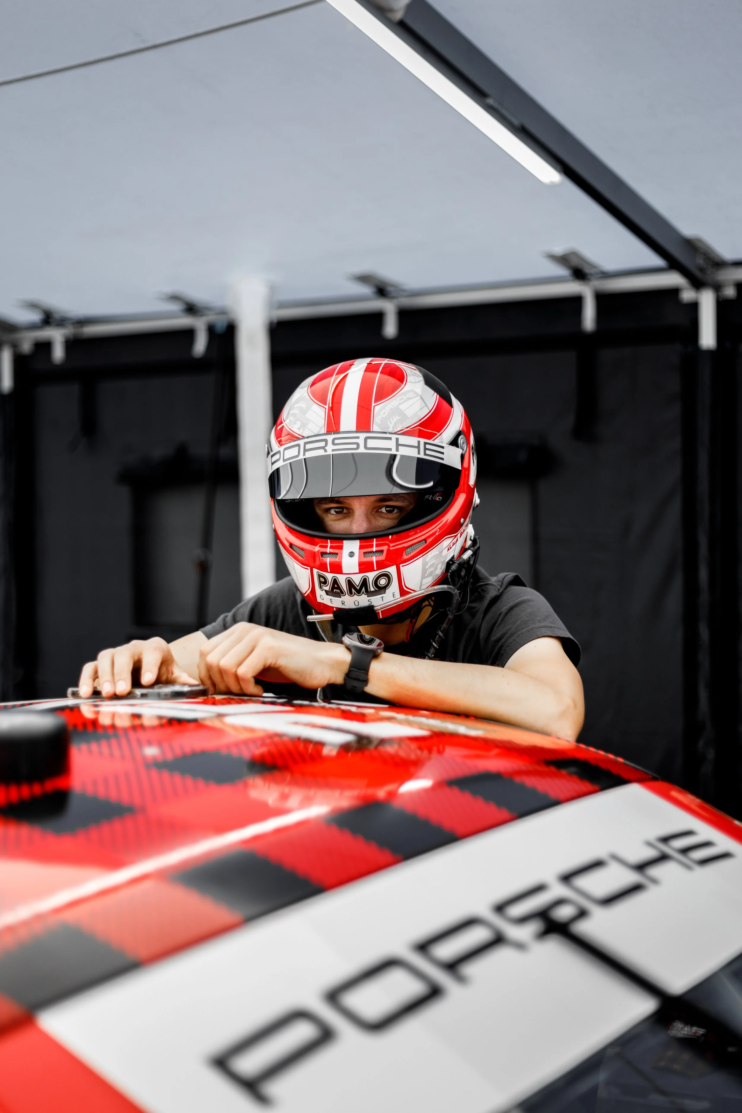 A race car driver wearing a red, black, and white helmet, leaning over a racing car with the Porsche logo, inside a garage or pit area.