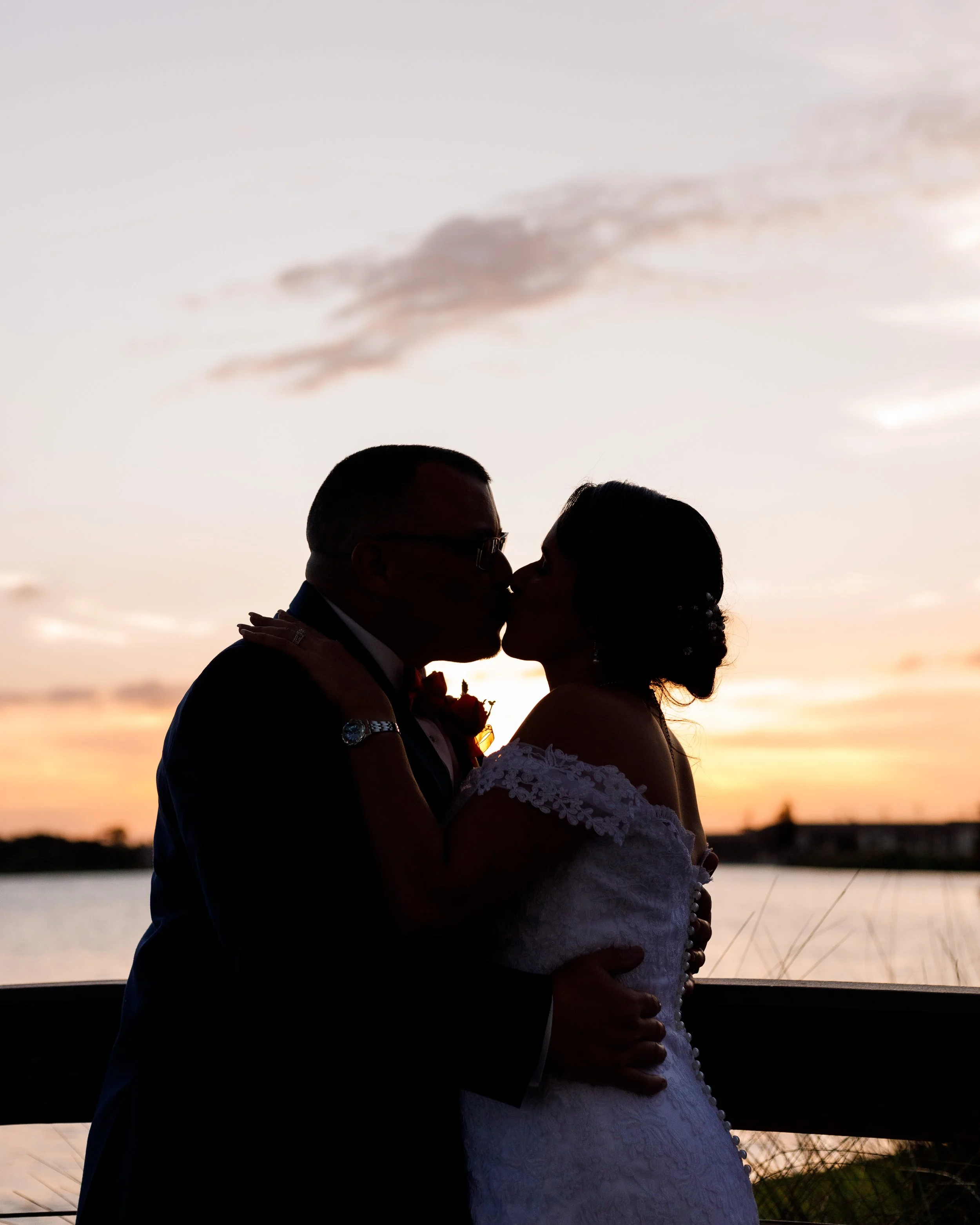Silhouette of a couple kissing by a lake at sunset, with a colorful sky and some clouds.