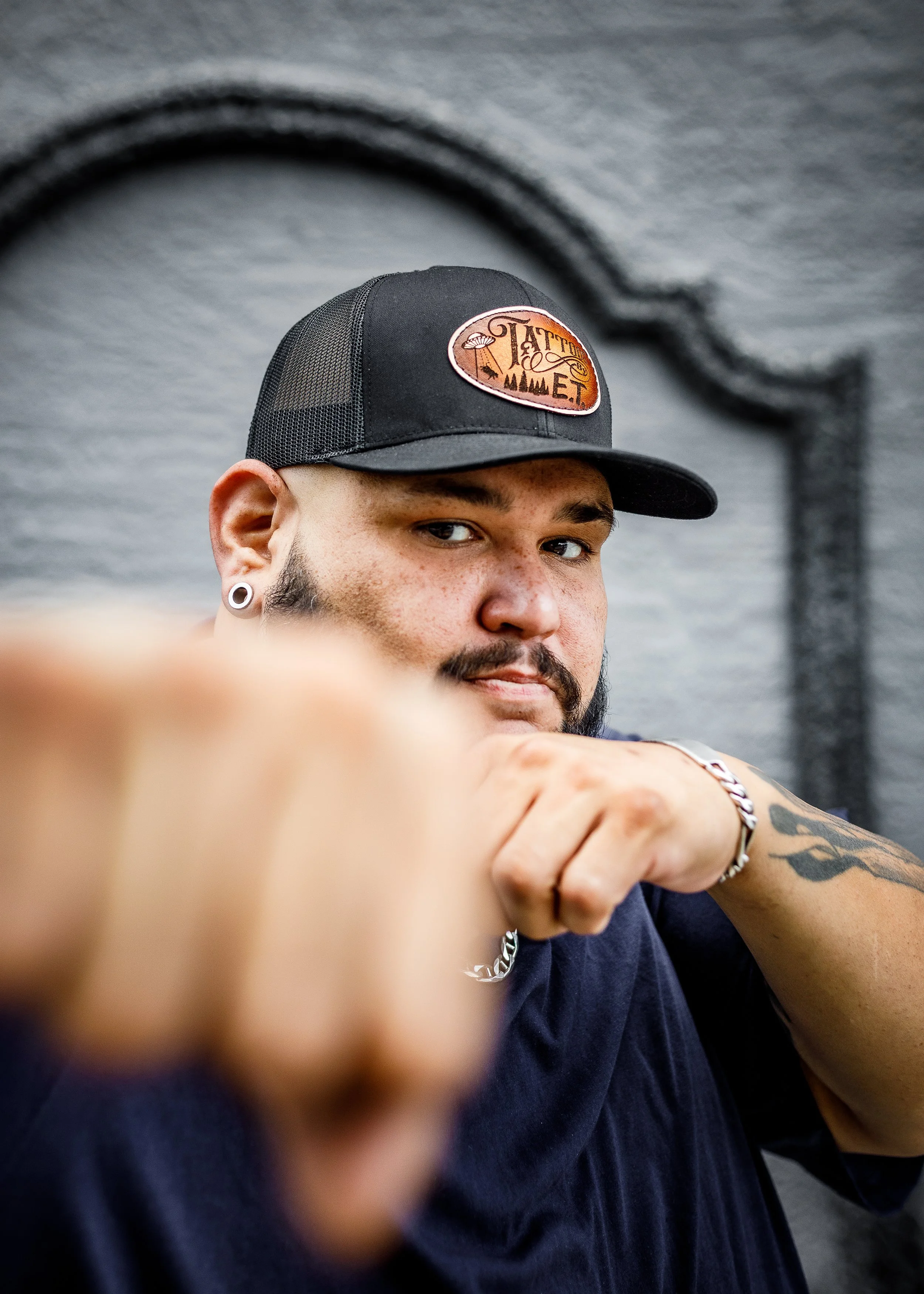 A man wearing a black trucker hat with a patch that says 'Tattoo Met,' pointing towards the camera with a confident expression, in front of a gray brick wall.