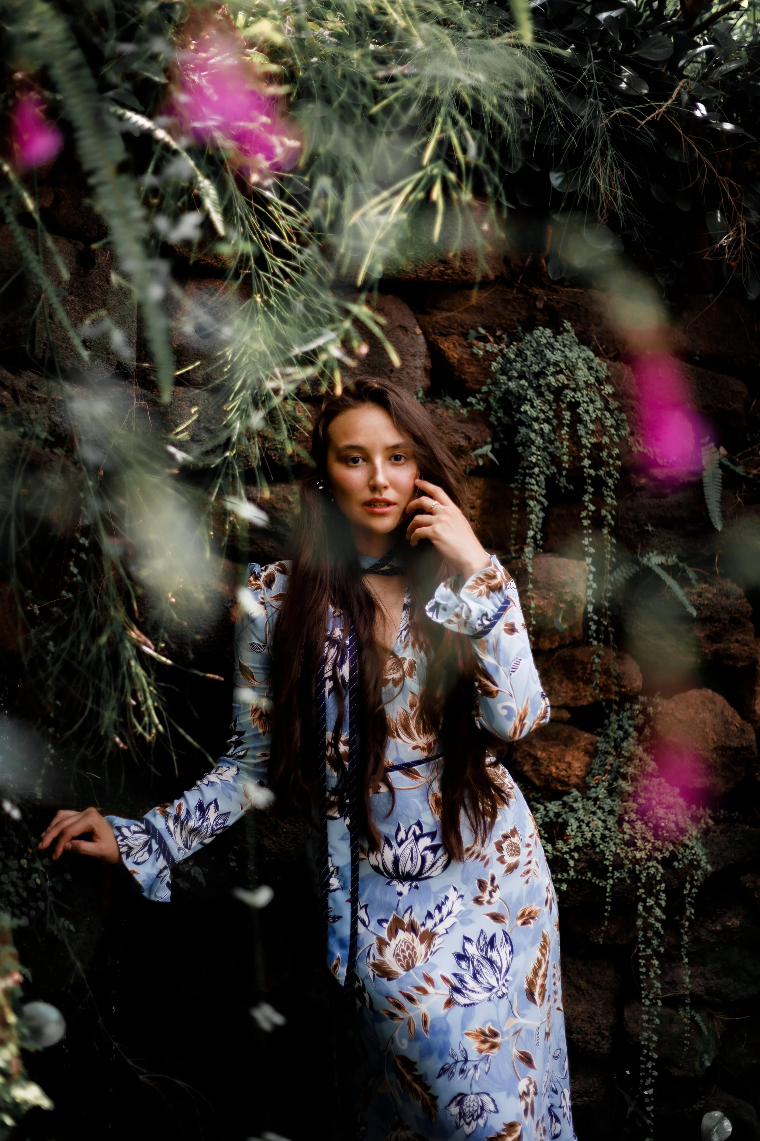 A woman with long dark hair standing in front of a stone wall with hanging greenery, wearing a long floral dress.