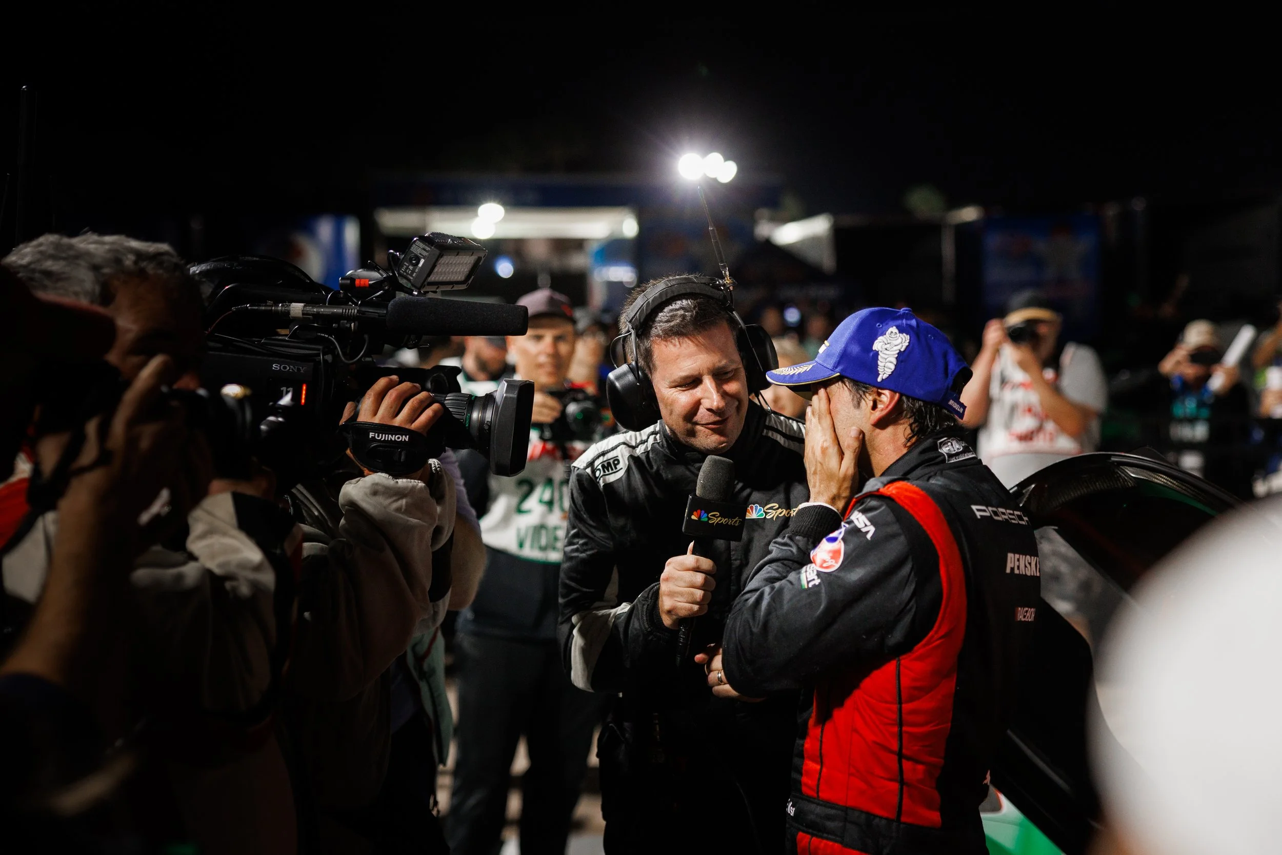 A race car driver in a black and red racing suit and blue cap covering his face with his hand, speaks to a man holding a microphone with the NBC Sports logo during a nighttime event, with a camera crew filming them and several people in the background.