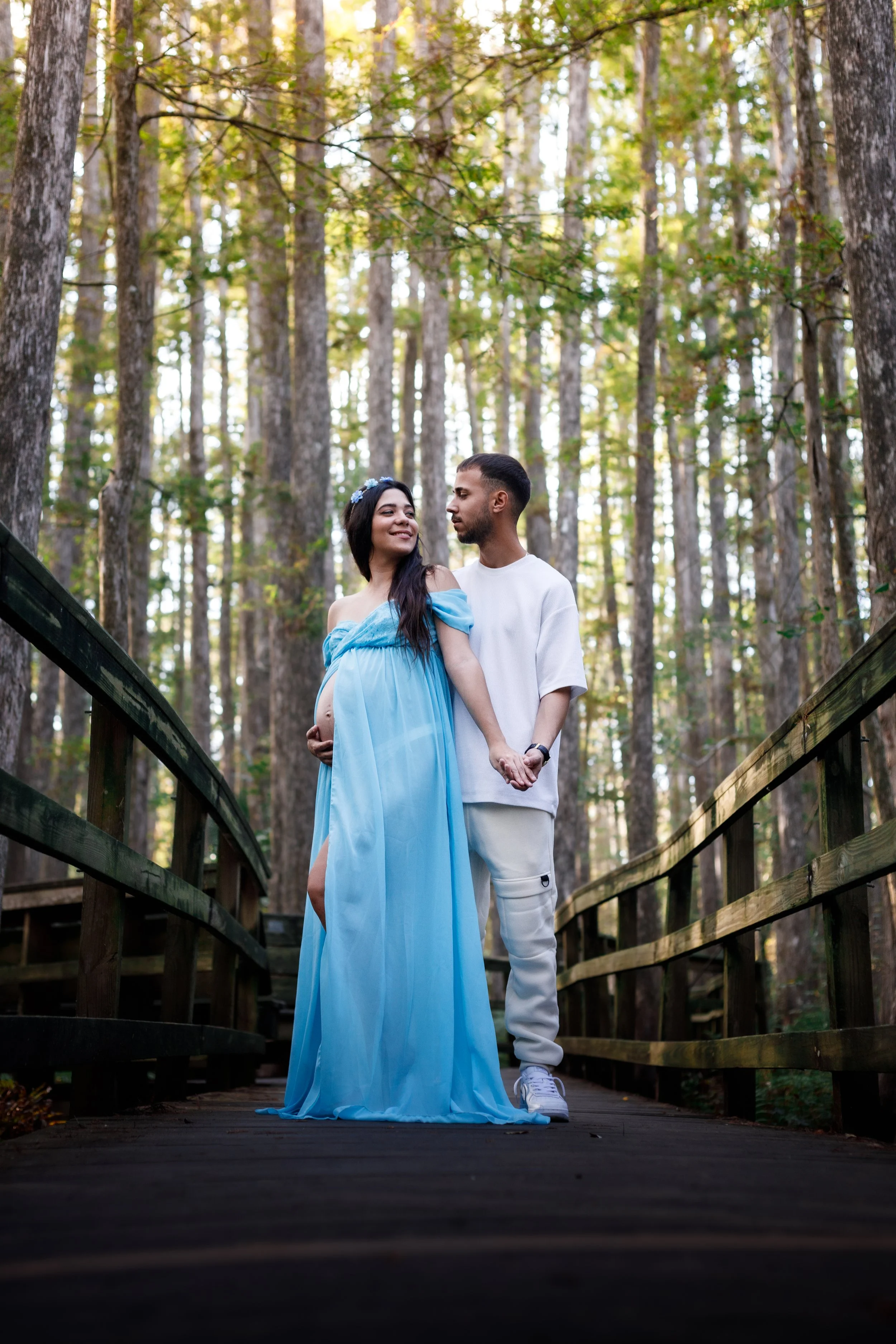 A pregnant woman and her partner standing on a wooden bridge in a forest, holding hands. The woman is wearing a blue, off-shoulder gown, and the man is in white casual clothing. The woman is smiling and has long dark hair, with trees and sunlight in 