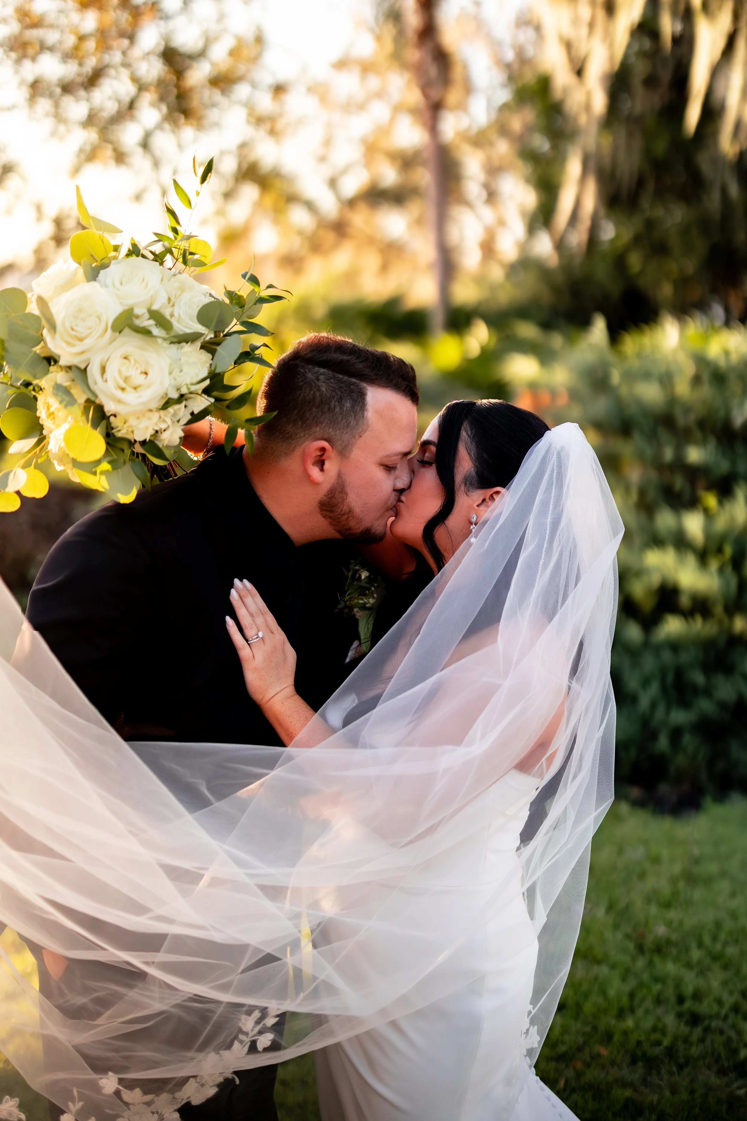 Bride and groom kissing outdoors, the bride's veil flowing, floral bouquet nearby, warm sunset lighting.