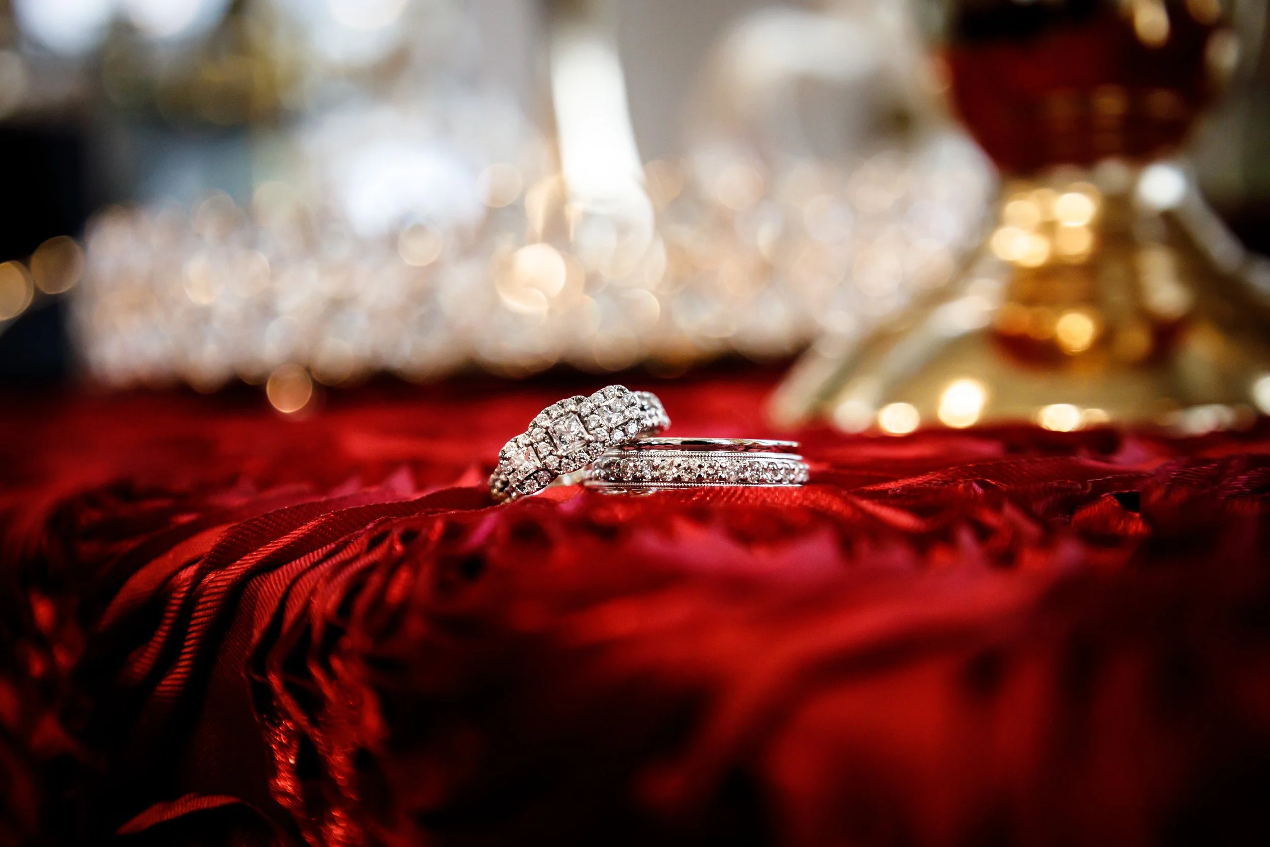 Close-up of two diamond rings on a red velvet fabric with blurred jewelry and gold objects in the background.