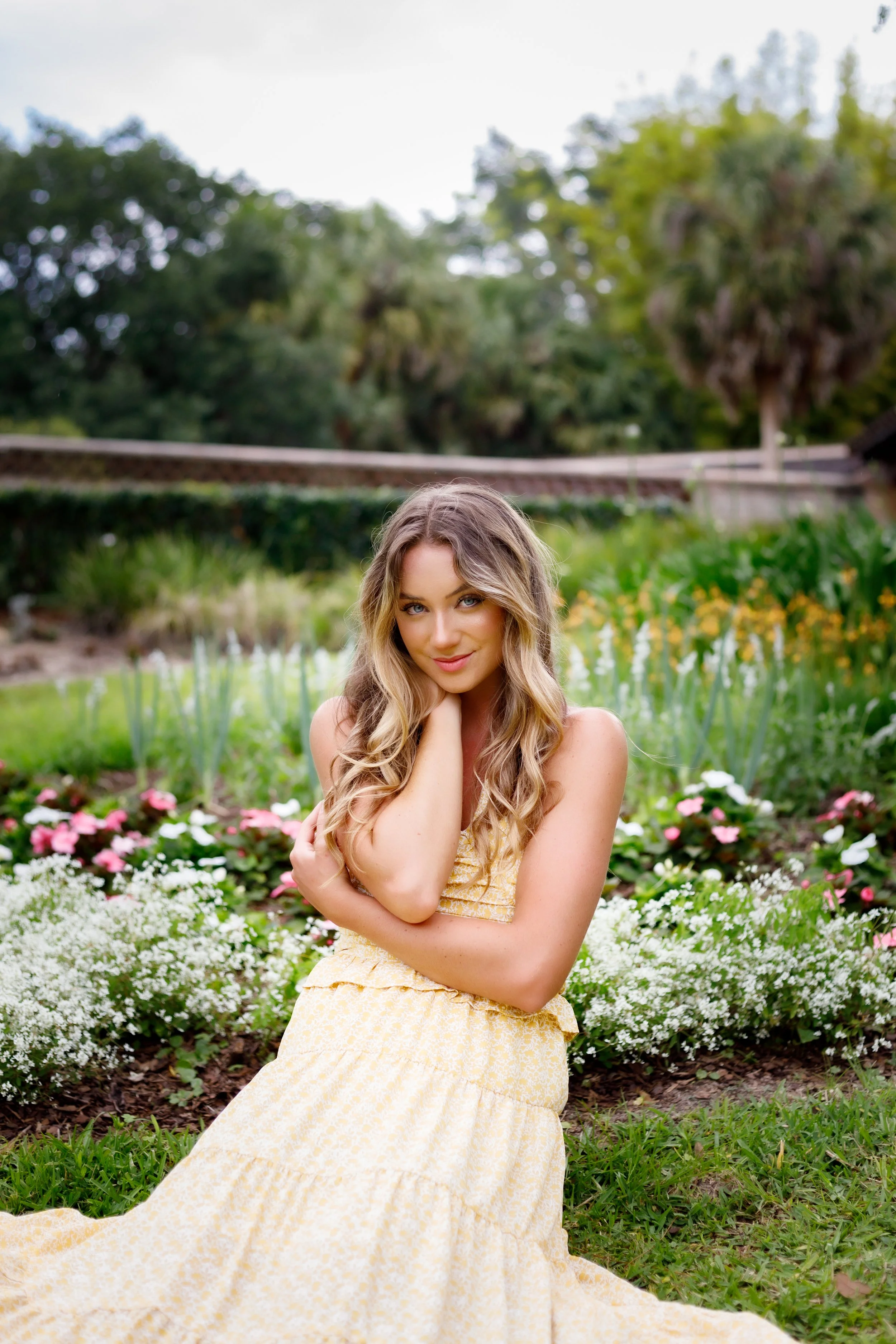 A woman with long wavy blonde hair and blue eyes sitting in a garden with pink and white flowers, wearing a yellow patterned dress, smiling and looking at the camera.