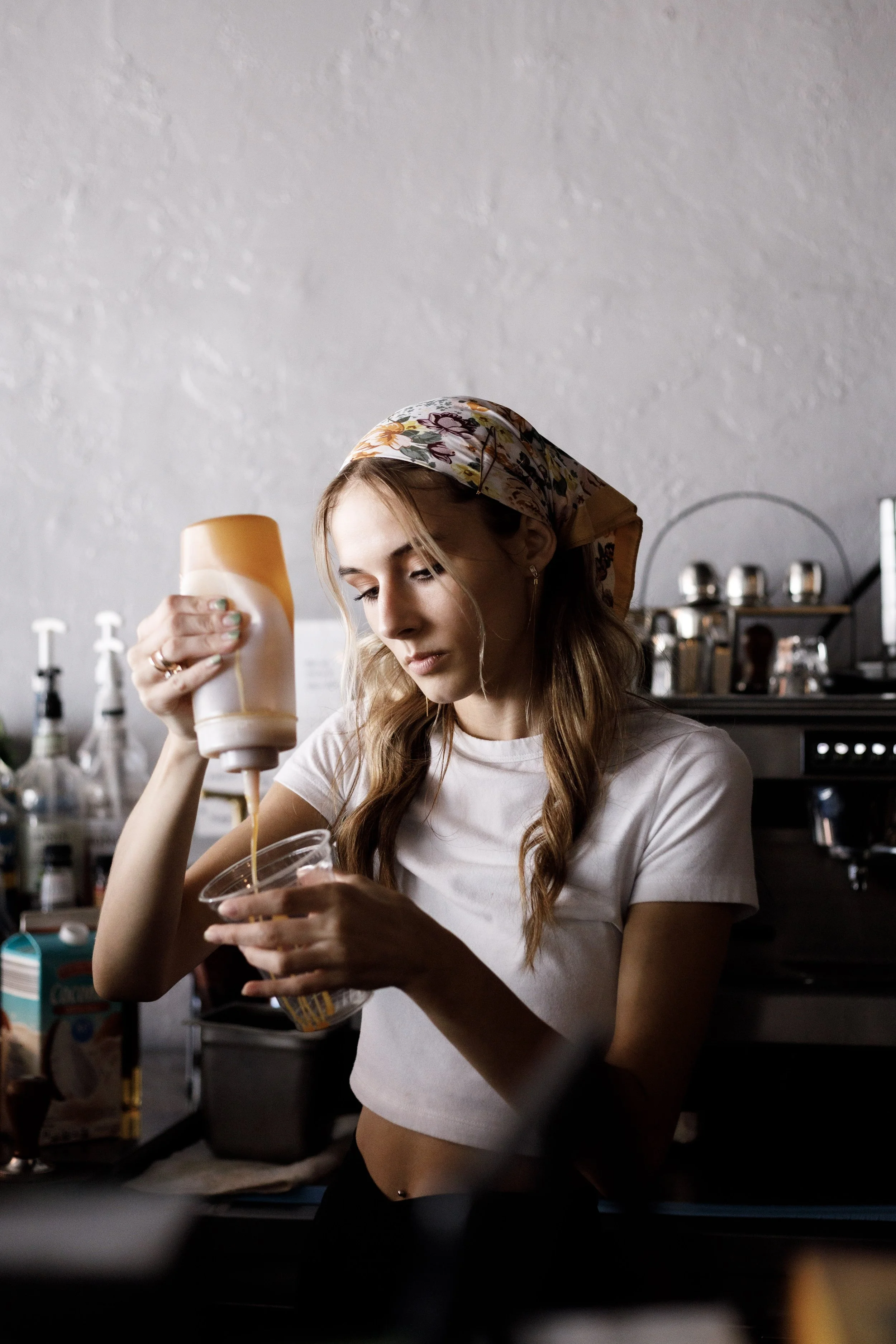 A young woman with a floral scarf on her head, wearing a white crop top, pouring ketchup from a bottle into a transparent container in a kitchen setting.