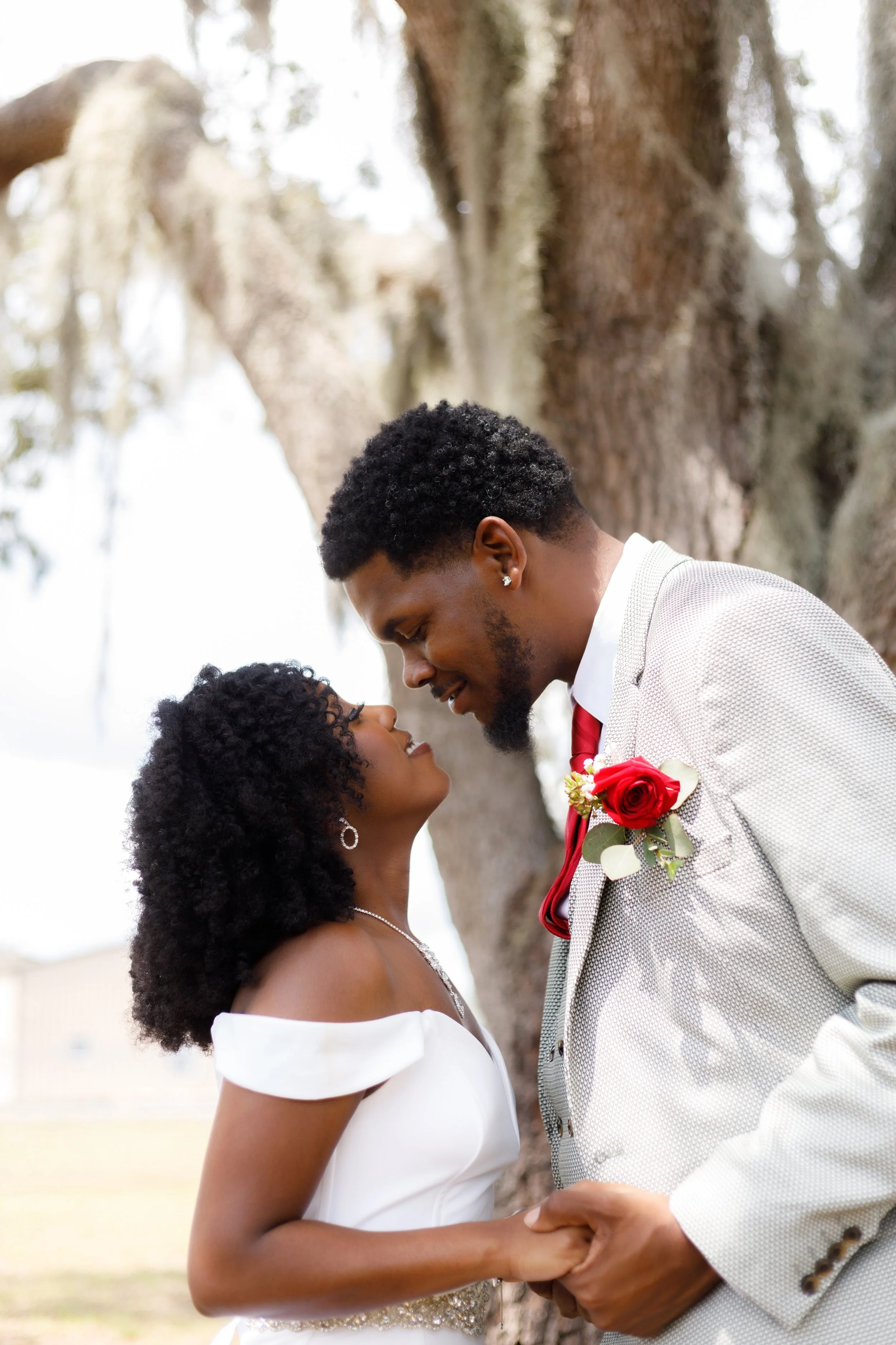 A wedding couple, an African-American man in a gray suit with a red boutonniere and a woman in a white off-the-shoulder dress with jewelry, share a moment under a large tree outdoors.