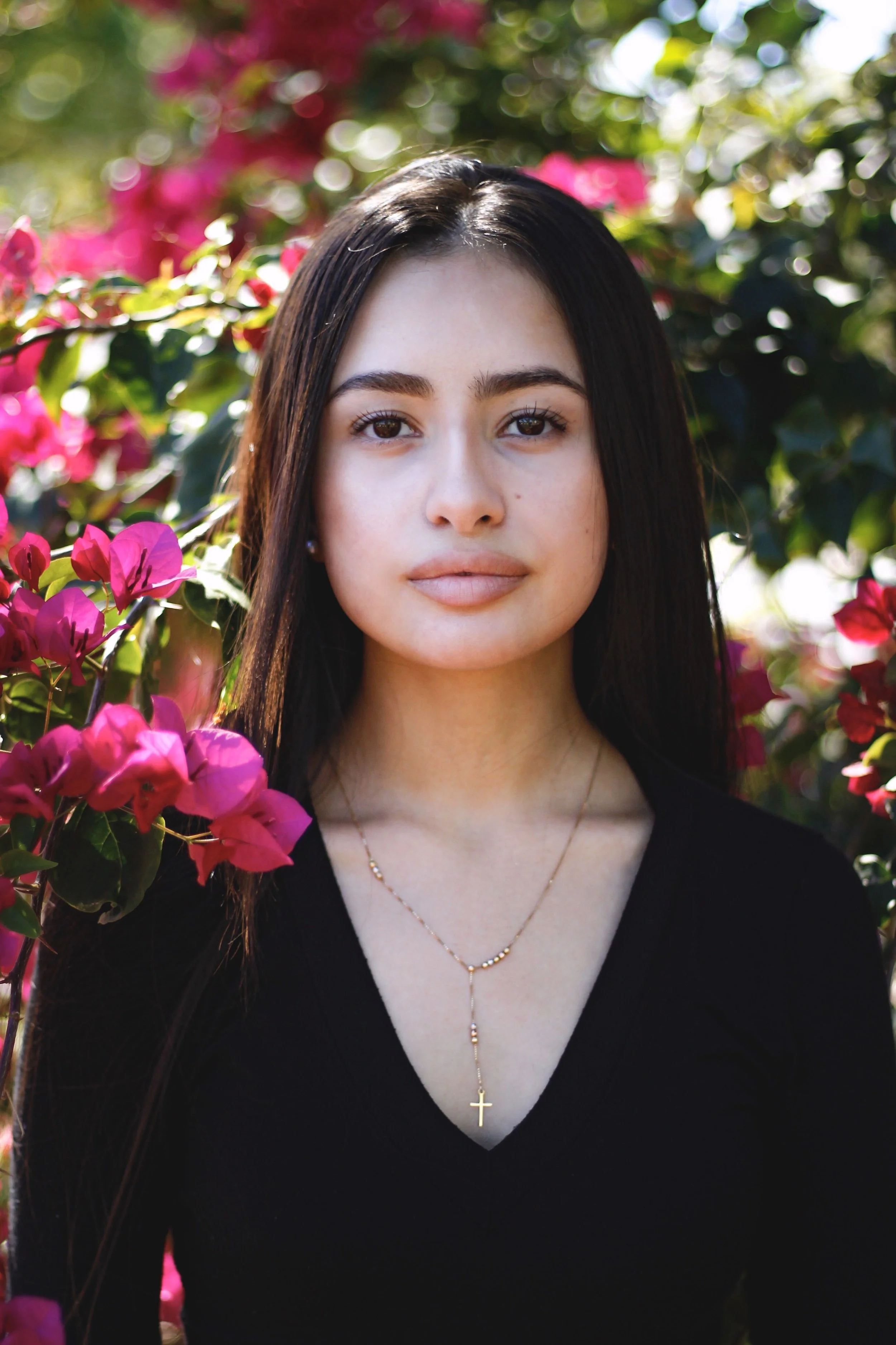 Portrait of a young woman with dark hair, wearing a black top and a gold cross necklace, standing outdoors among pink flowers and green foliage.