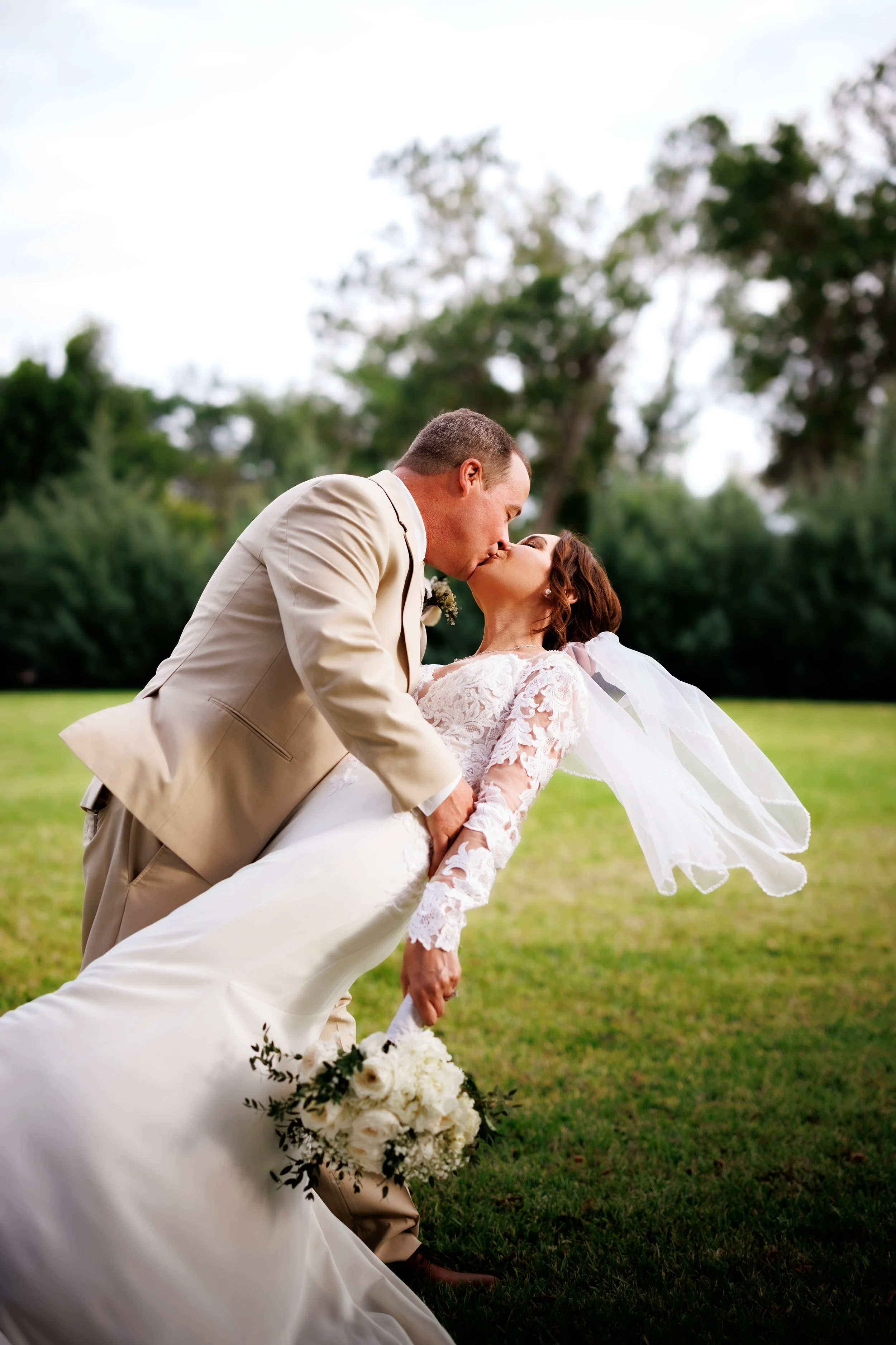 A newlywed couple sharing a kiss outdoors, with the groom holding the bride as she leans back holding a bouquet of white flowers, in a grassy area with trees in the background.