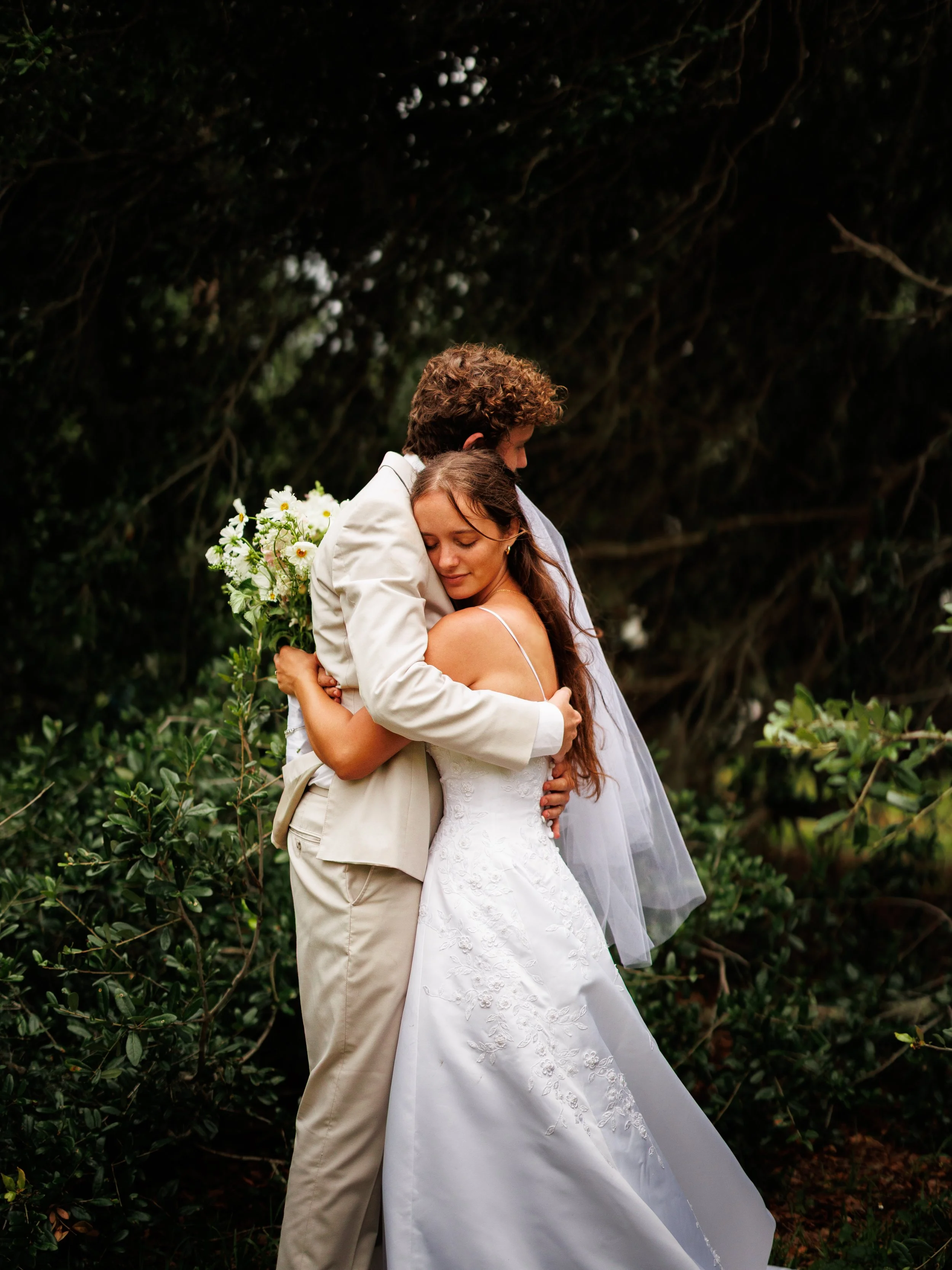 A couple dressed in wedding attire sharing a hug outdoors, surrounded by greenery.