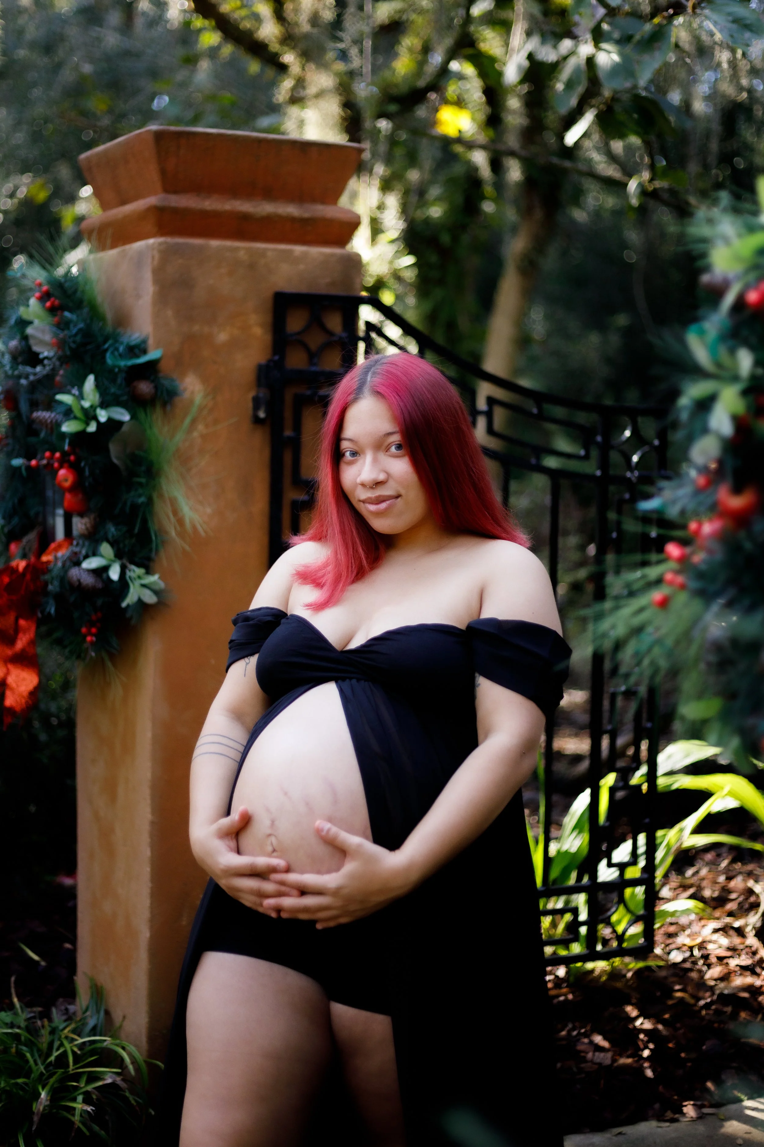 Pregnant woman with pink hair standing outdoors near a decorated fence, holding her belly with both hands.