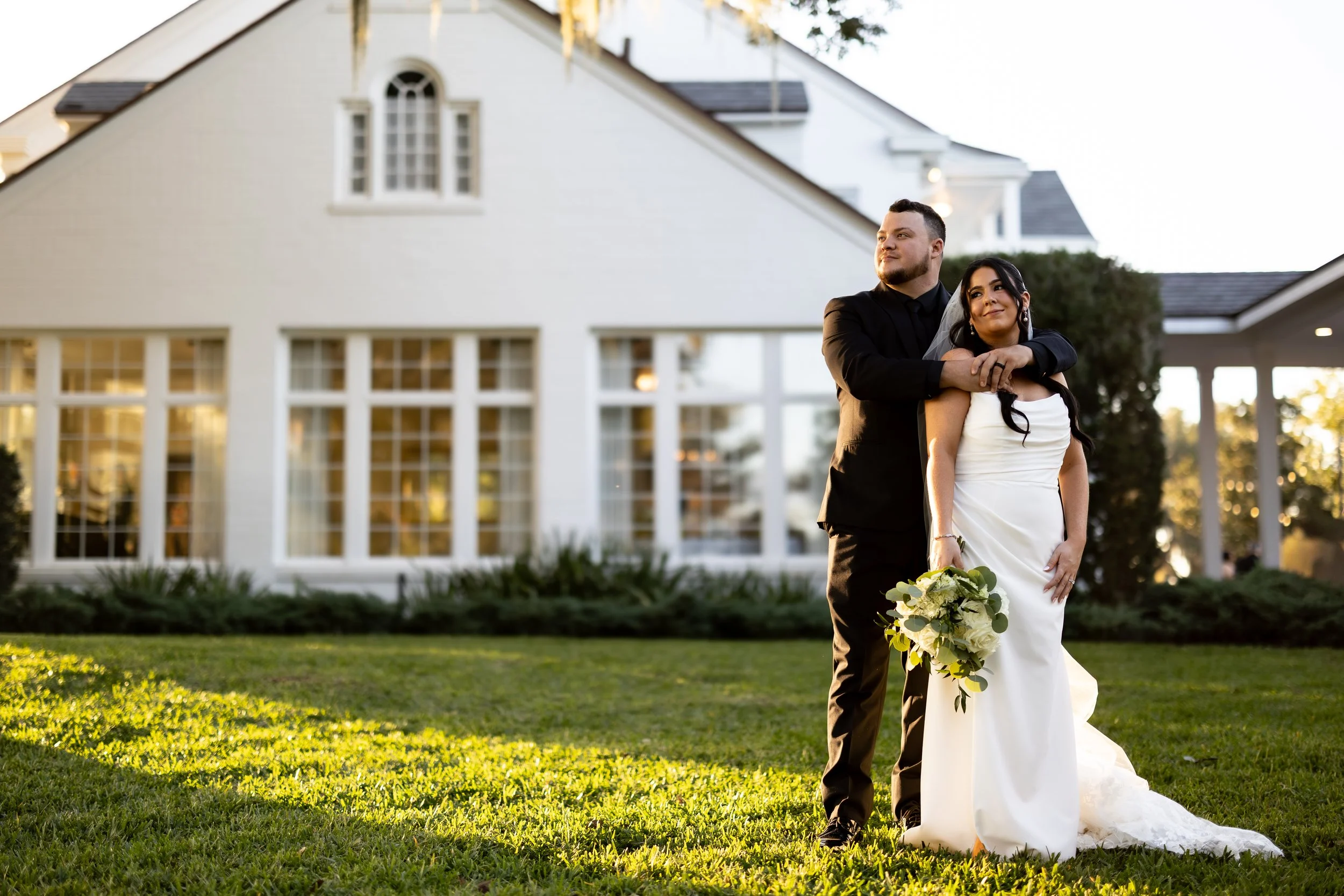 A newlywed couple standing on a lawn in front of a large white house during sunset. The groom, dressed in a black suit, gently holds the bride from behind. The bride, in a white wedding gown, holds a bouquet of white flowers and gazes thoughtfully into the distance.