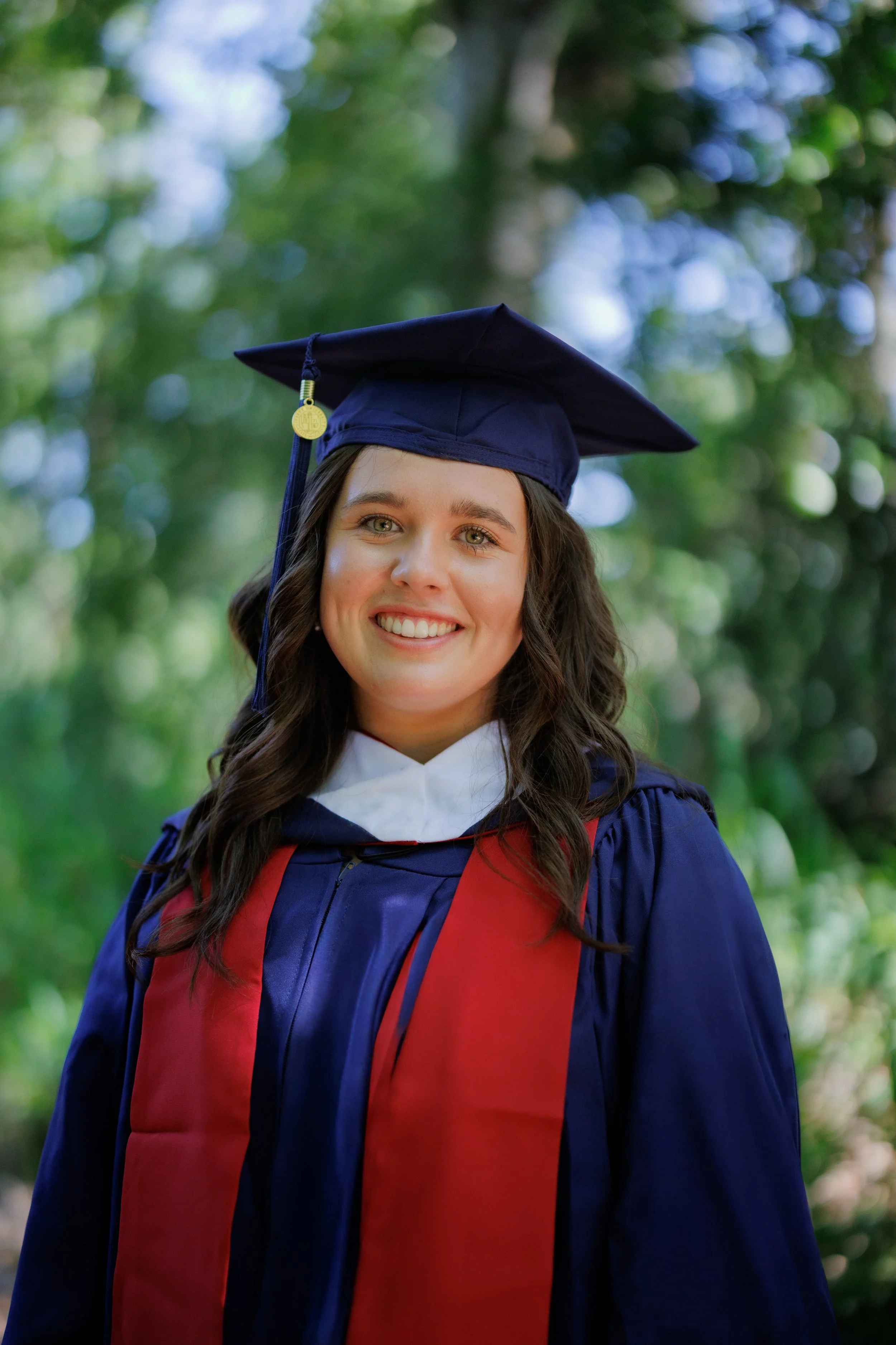 A young woman in a blue graduation gown and cap with a red stole, standing outdoors with a blurred green foliage background, smiling.