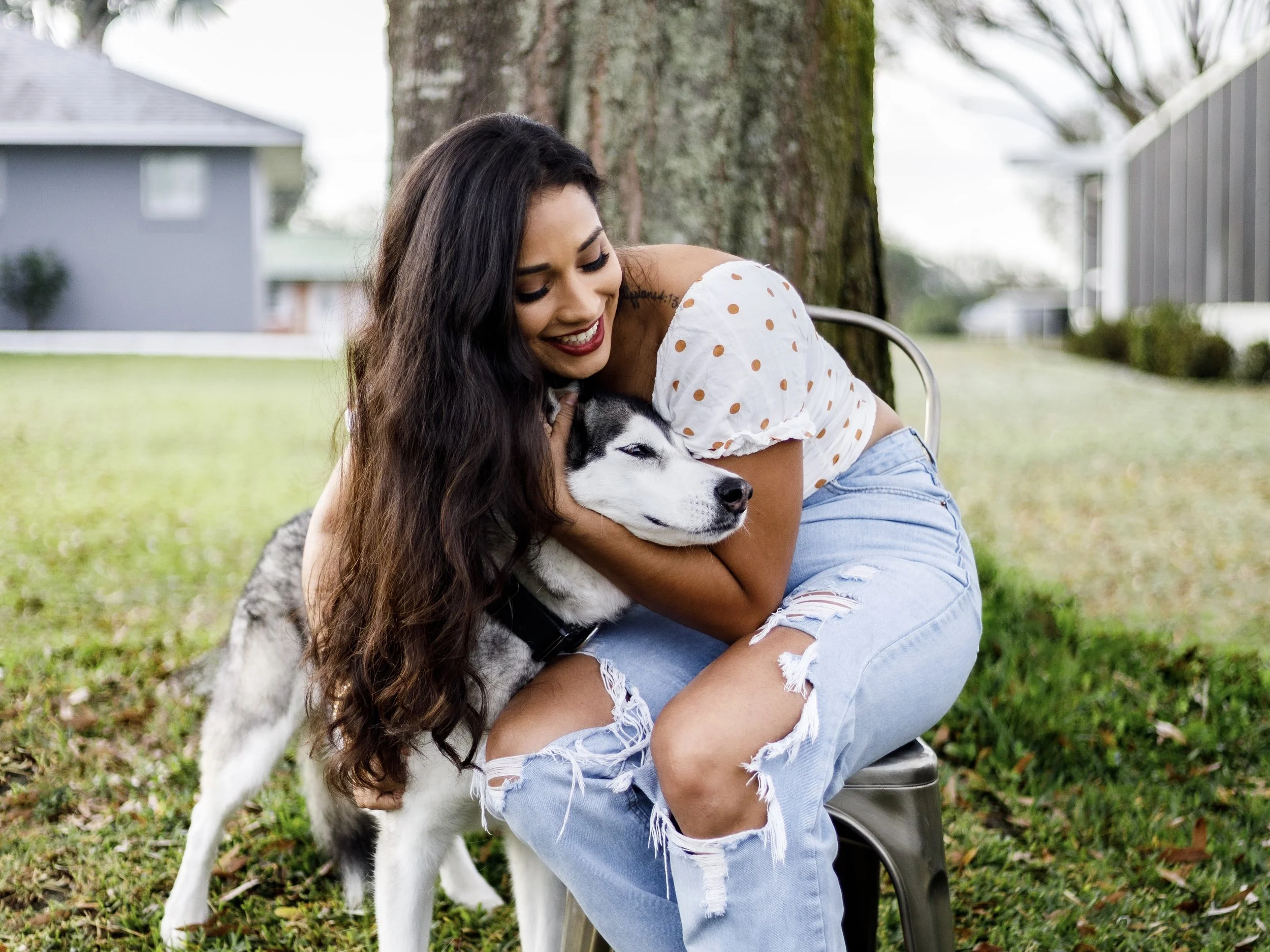 A woman with long brown hair, wearing a white polka dot top and ripped jeans, sitting outdoors hugging a Siberian Husky puppy.